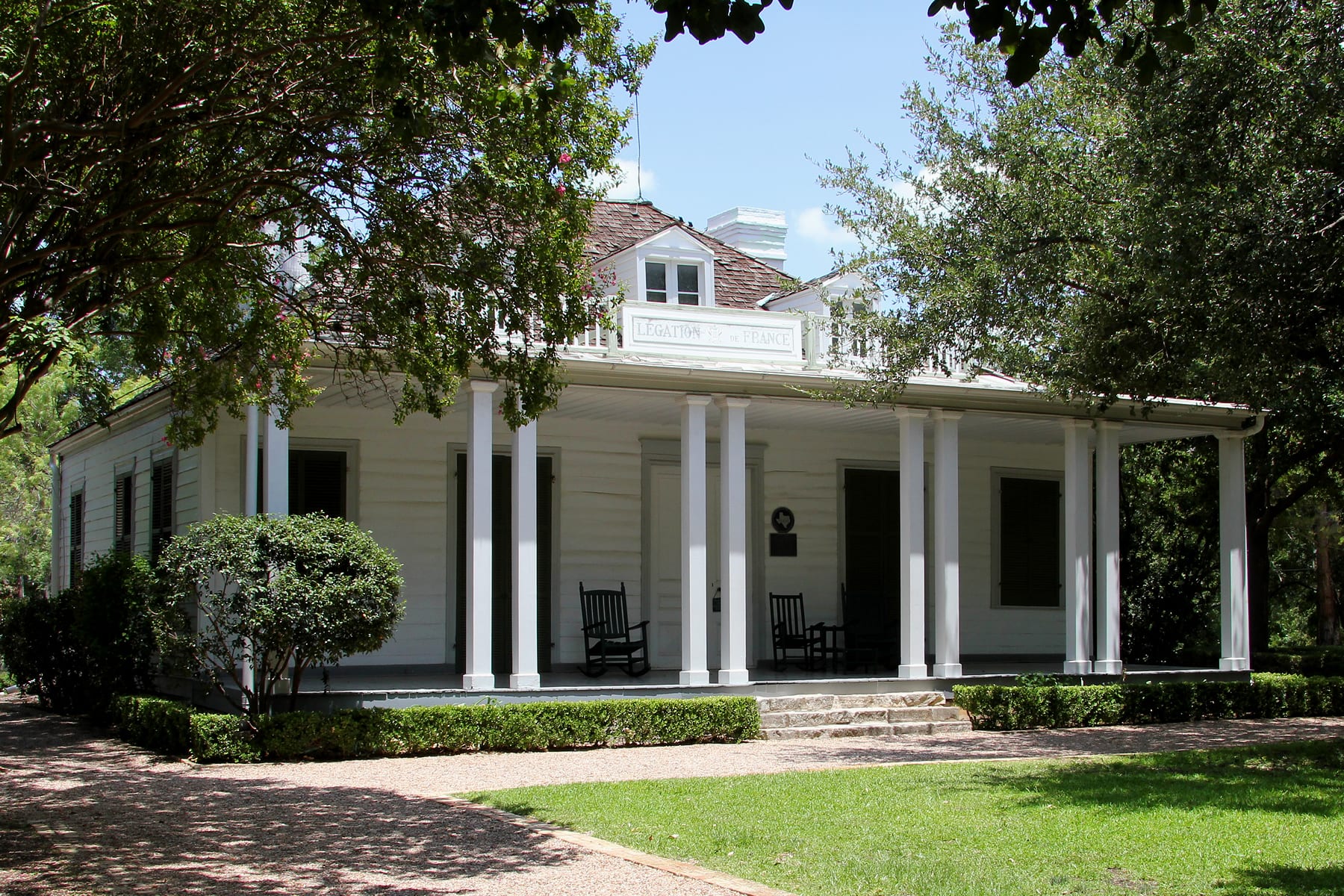 A white single-storey wooden house with a deep columned porch and a sign reading Légation de France, shaded by trees