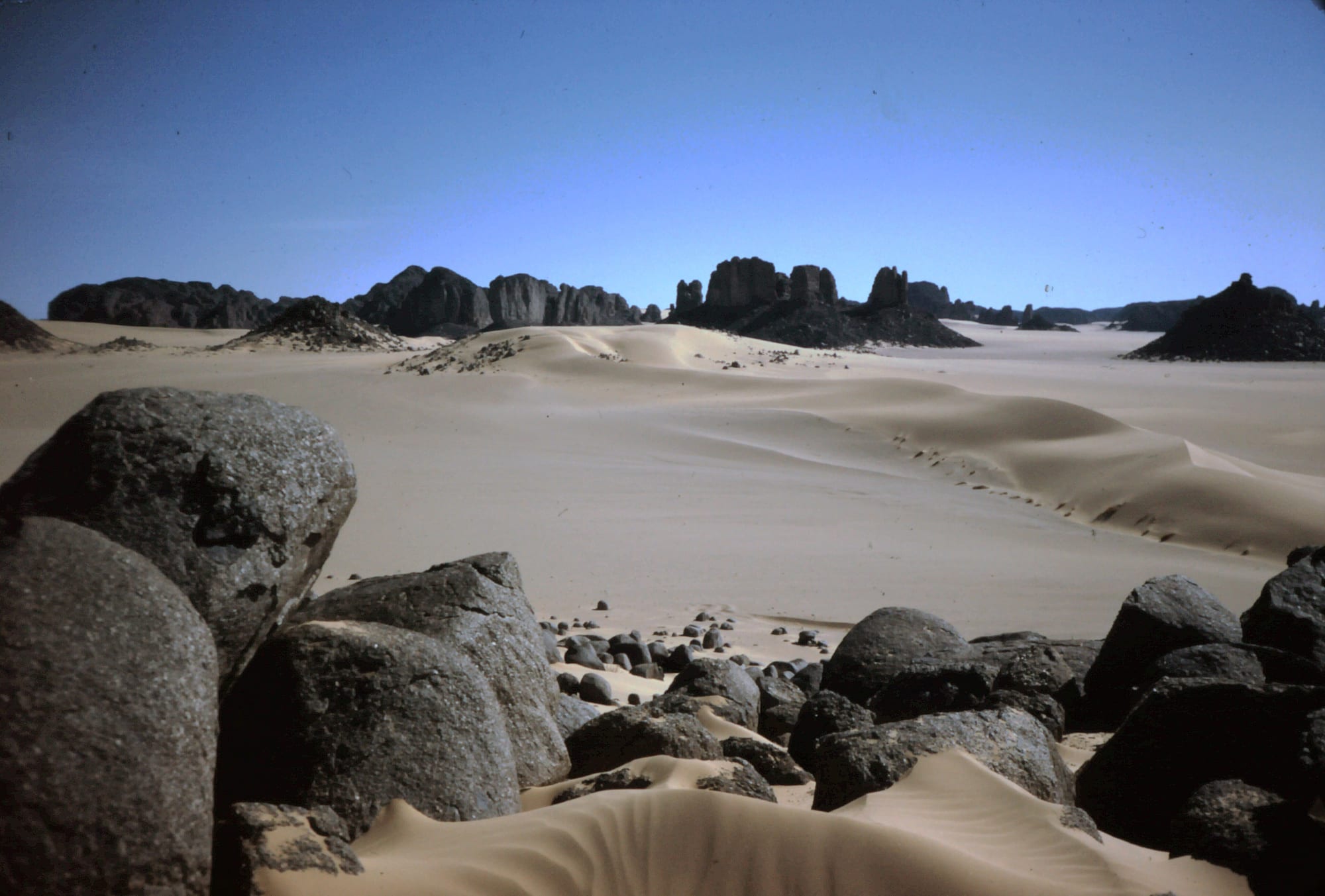 Pale sand dunes between dark rounded boulders, with eroded black rock towers on the horizon under a deep blue sky