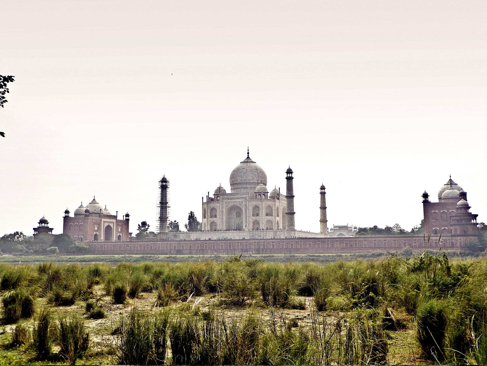 A white domed tomb flanked by two red sandstone buildings with domes, behind a long wall, seen across grassland