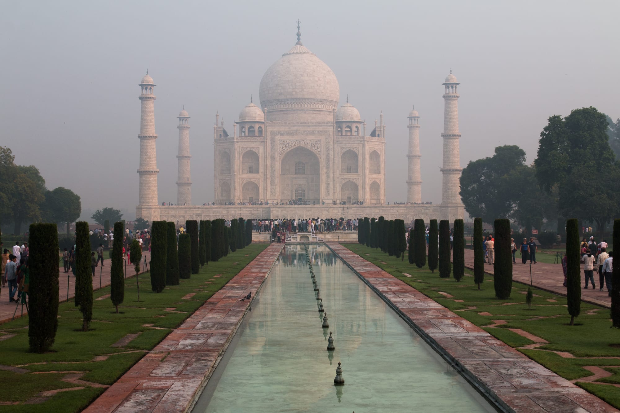 A white marble domed mausoleum with four minarets at the end of a long reflecting pool lined with cypress trees