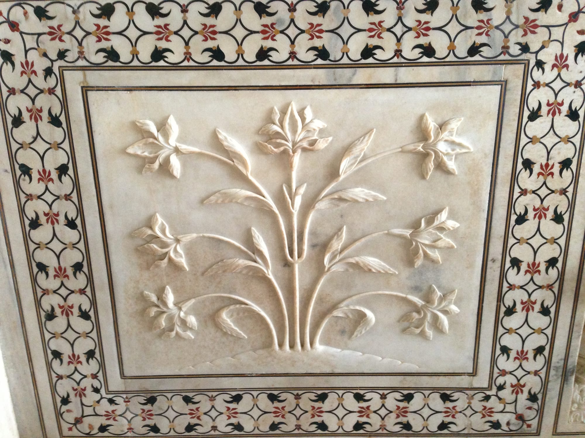 A white marble panel carved in relief with a flowering plant, framed by a border of inlaid red and black stone flowers and leaves