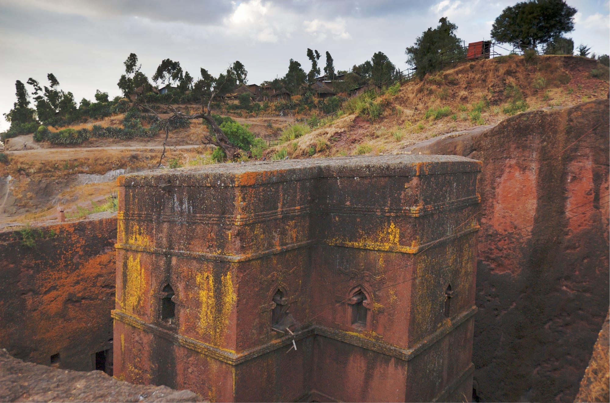 A tall cross-shaped church of reddish rock streaked with yellow lichen, standing in a deep pit with sheer rock walls and a hillside of trees above