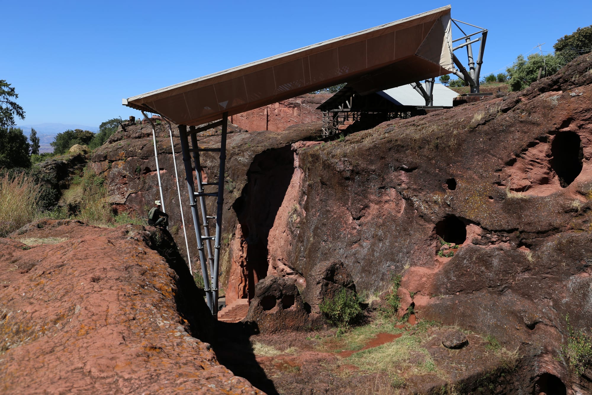 A deep, narrow trench cut through red rock, with a metal shelter roof on steel supports above the church beyond