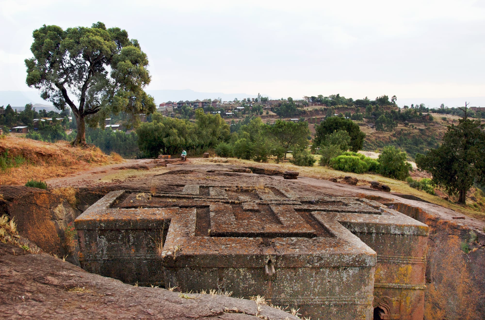 The flat roof of a church carved with a raised cross pattern, sitting level with the surrounding ground at the bottom of a rock pit, with trees and a hillside town behind