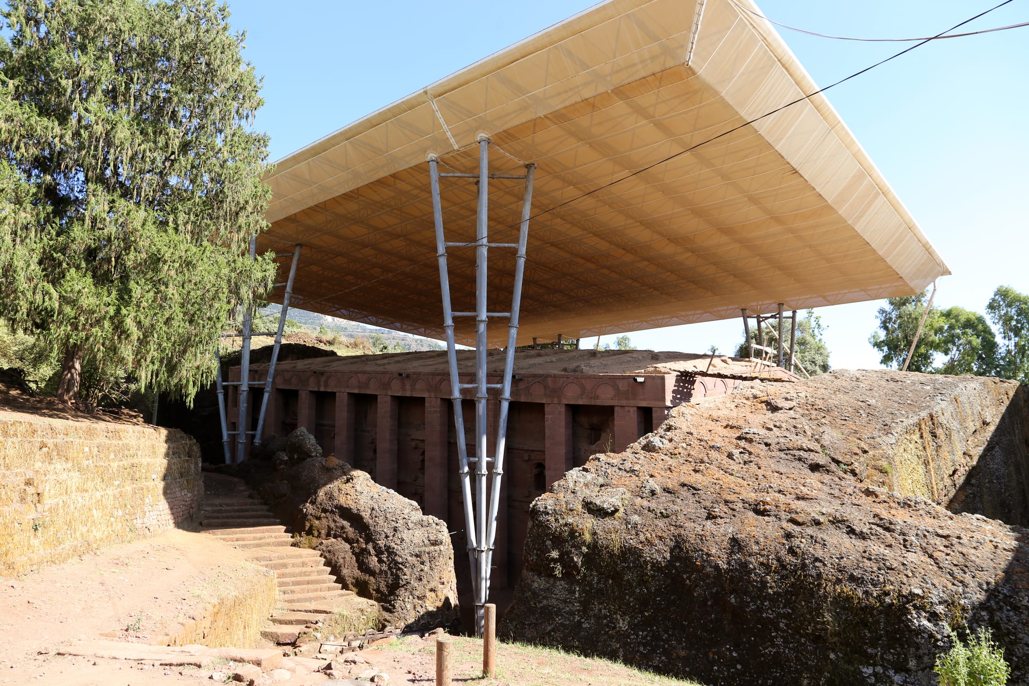 A large rock-cut church with a row of square pillars along its side, standing in a pit under a huge white protective canopy held up on steel legs