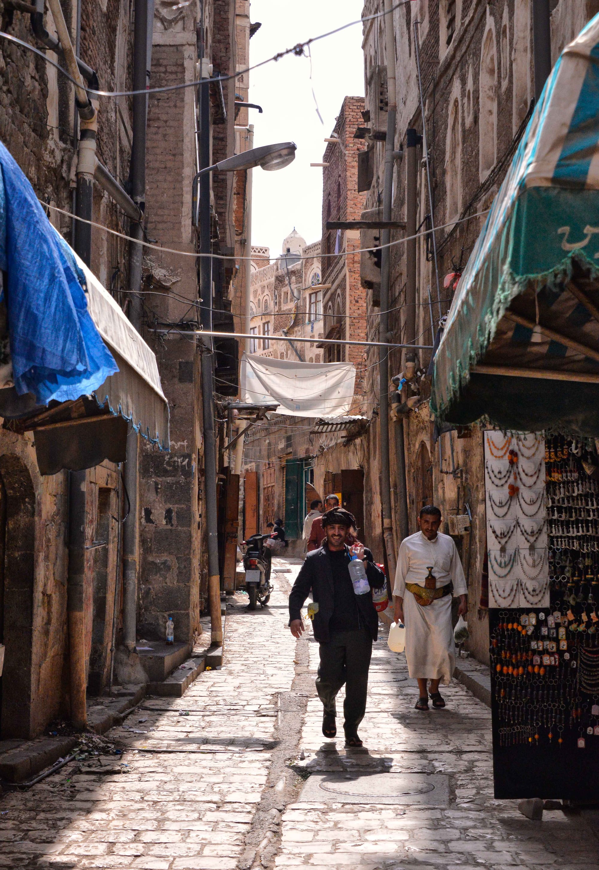 A narrow alley in Old Sana'a lined with decorated tower-houses and market stalls, a man carrying water bottles past a jewellery stand