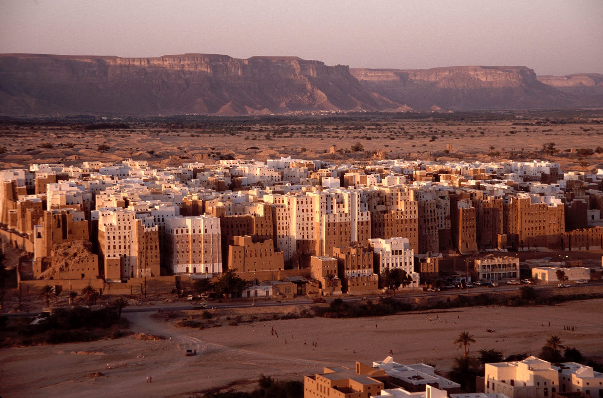 A dense cluster of tall mud-brick tower buildings rising from a desert valley