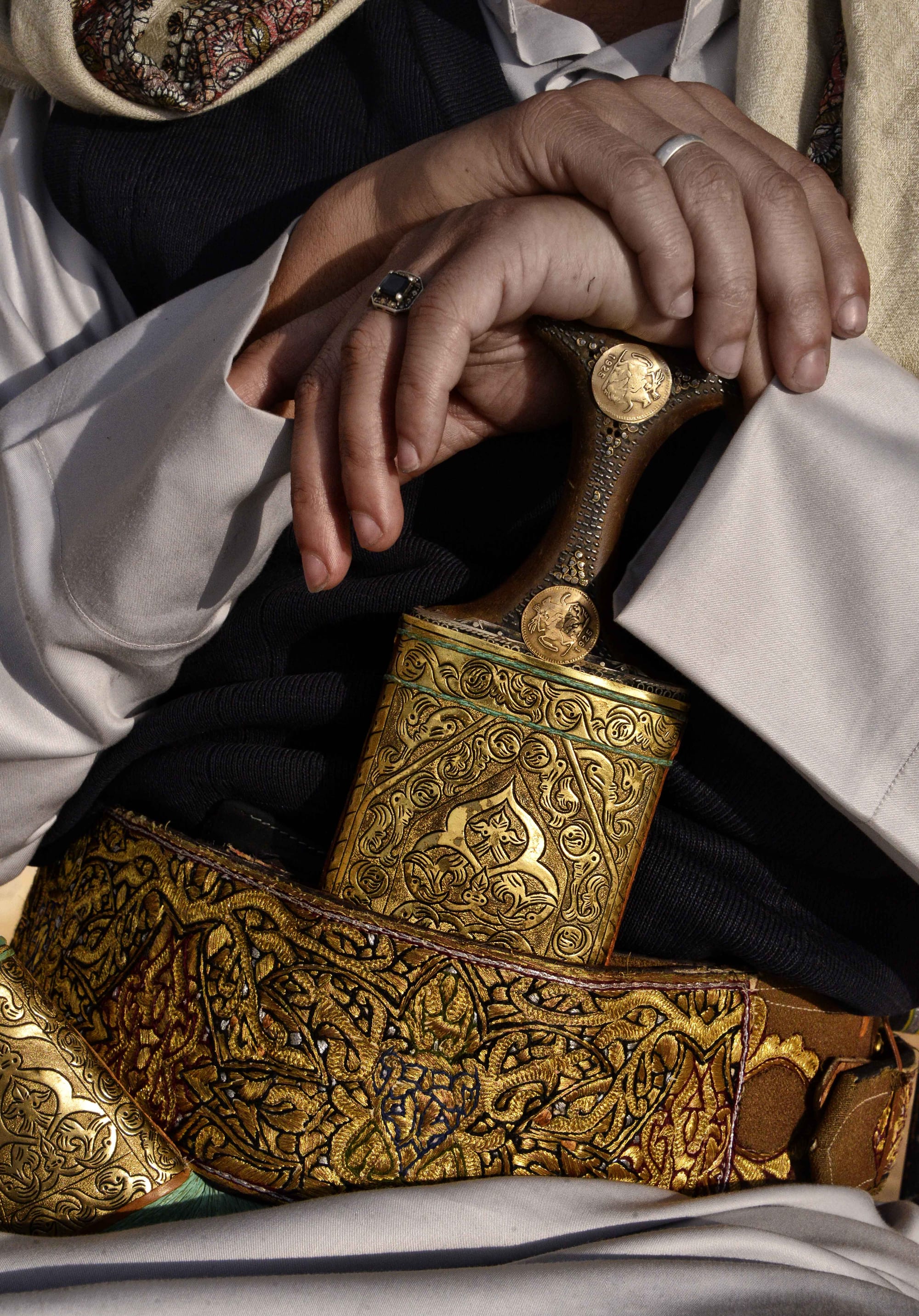 A man's hands resting on the dark studded hilt of a curved dagger in an engraved brass sheath, tucked into a wide belt embroidered with gold thread over a white robe