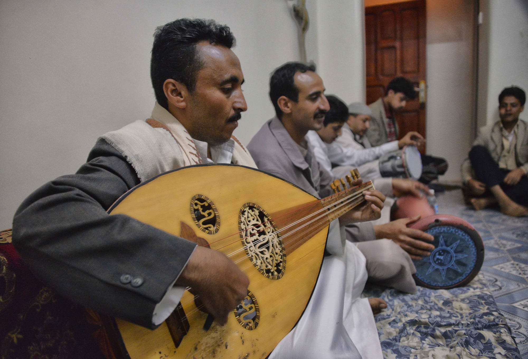 A man in a grey jacket and white shawl plays a large oud with an ornately carved soundboard, with other seated men playing drums behind him on a patterned carpet