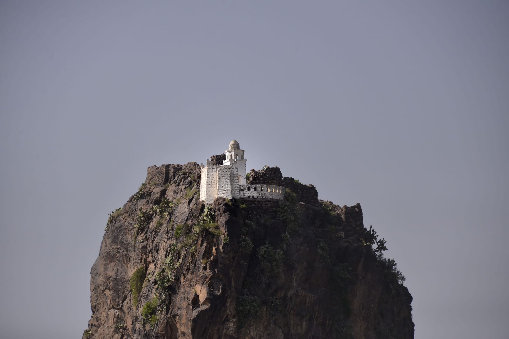 A small white mosque with a domed minaret stands on the summit of a steep, rugged rock outcrop against a grey sky