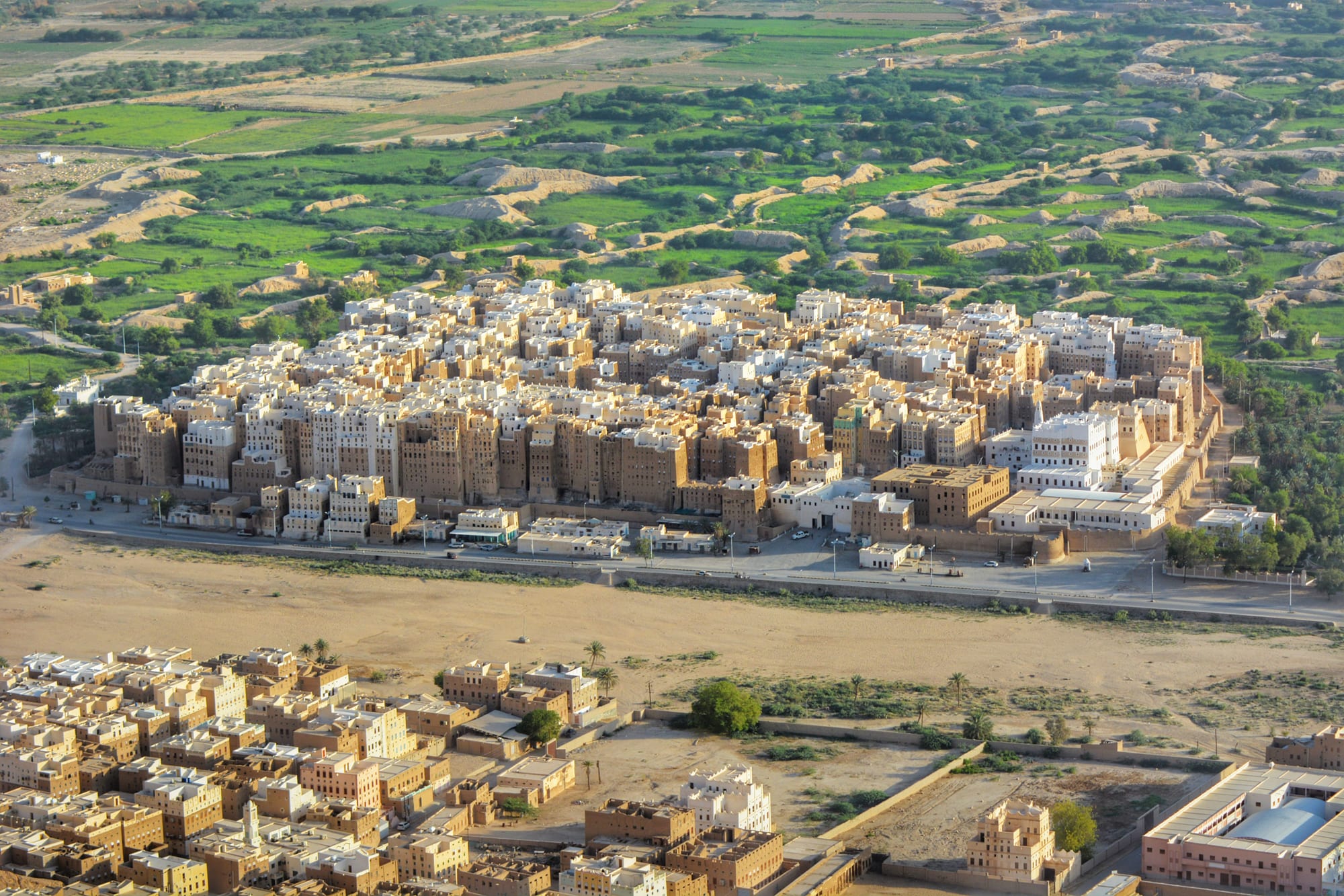 An aerial view of a dense rectangular cluster of tall pale mud-brick tower houses surrounded by a wall, set in a green valley with a newer town in the foreground