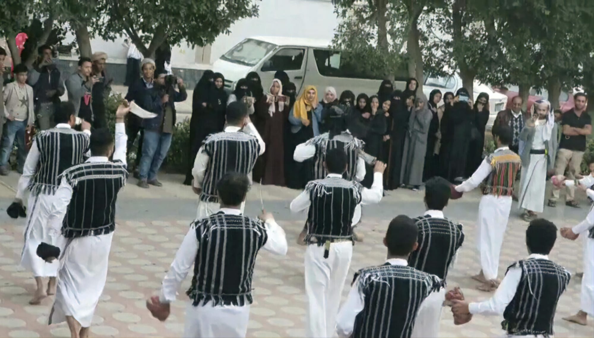 A group of men in long white robes and black waistcoats with white stripes dance in a circle on a paved square, watched by a crowd of men and women behind them