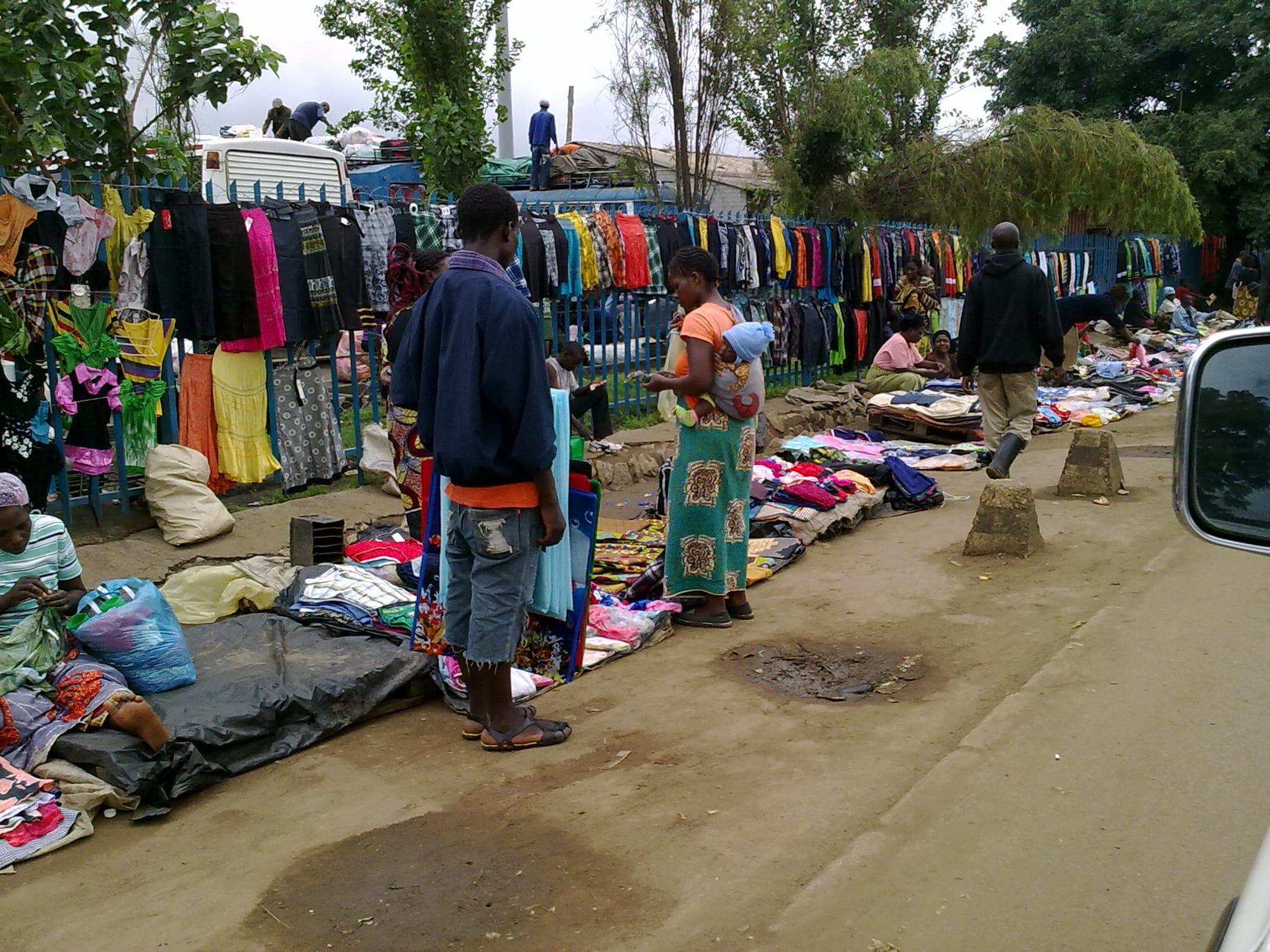 Vendors and shoppers at an outdoor market in Lusaka, with secondhand clothes hung along a fence and laid out on the ground