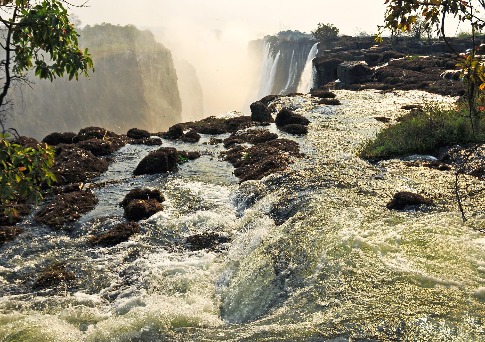 A wide river rushing between dark boulders towards the lip of a huge waterfall, with mist rising from the gorge beyond