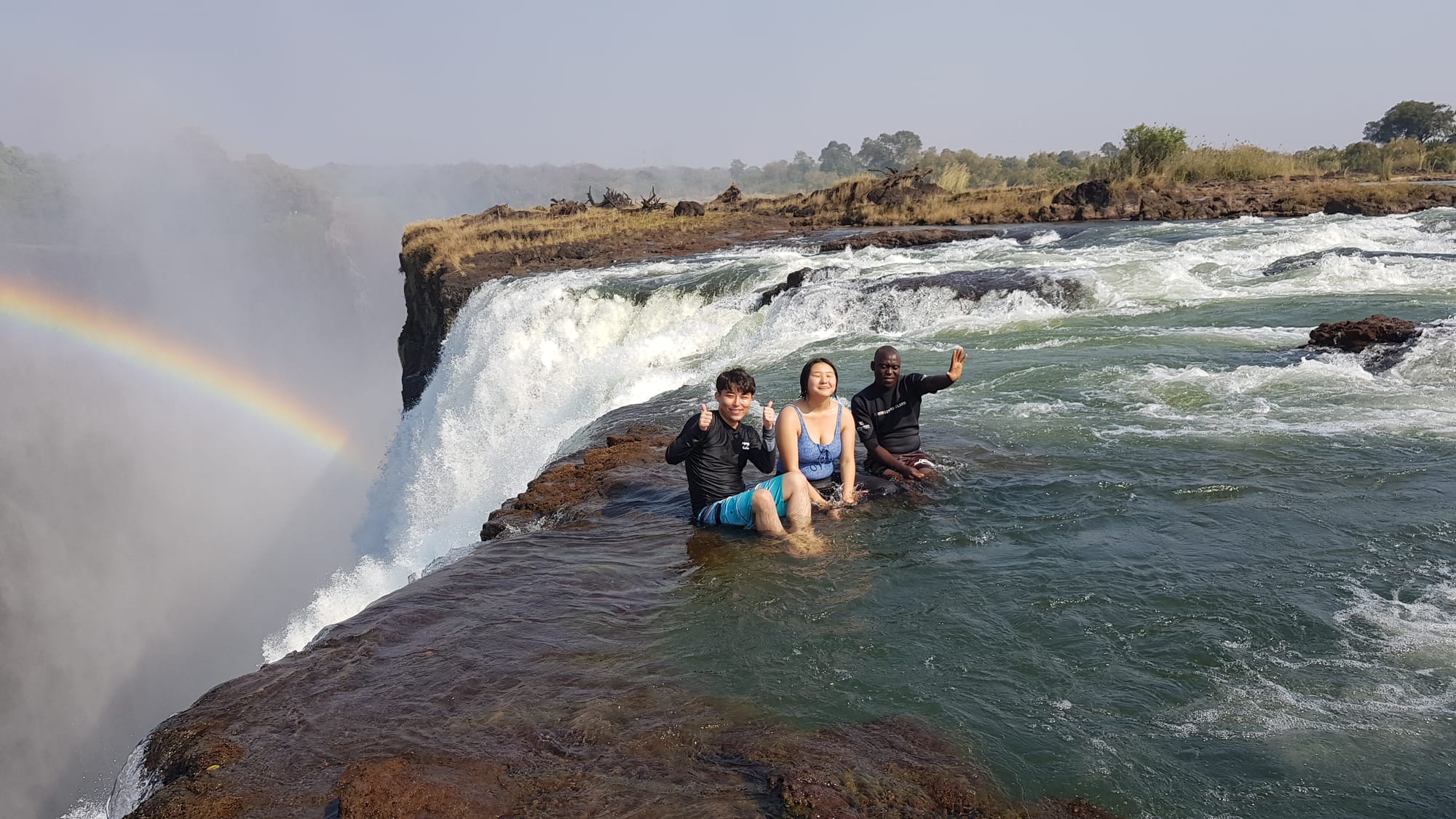 People swimming at the very edge of a waterfall, held back by a natural rock lip
