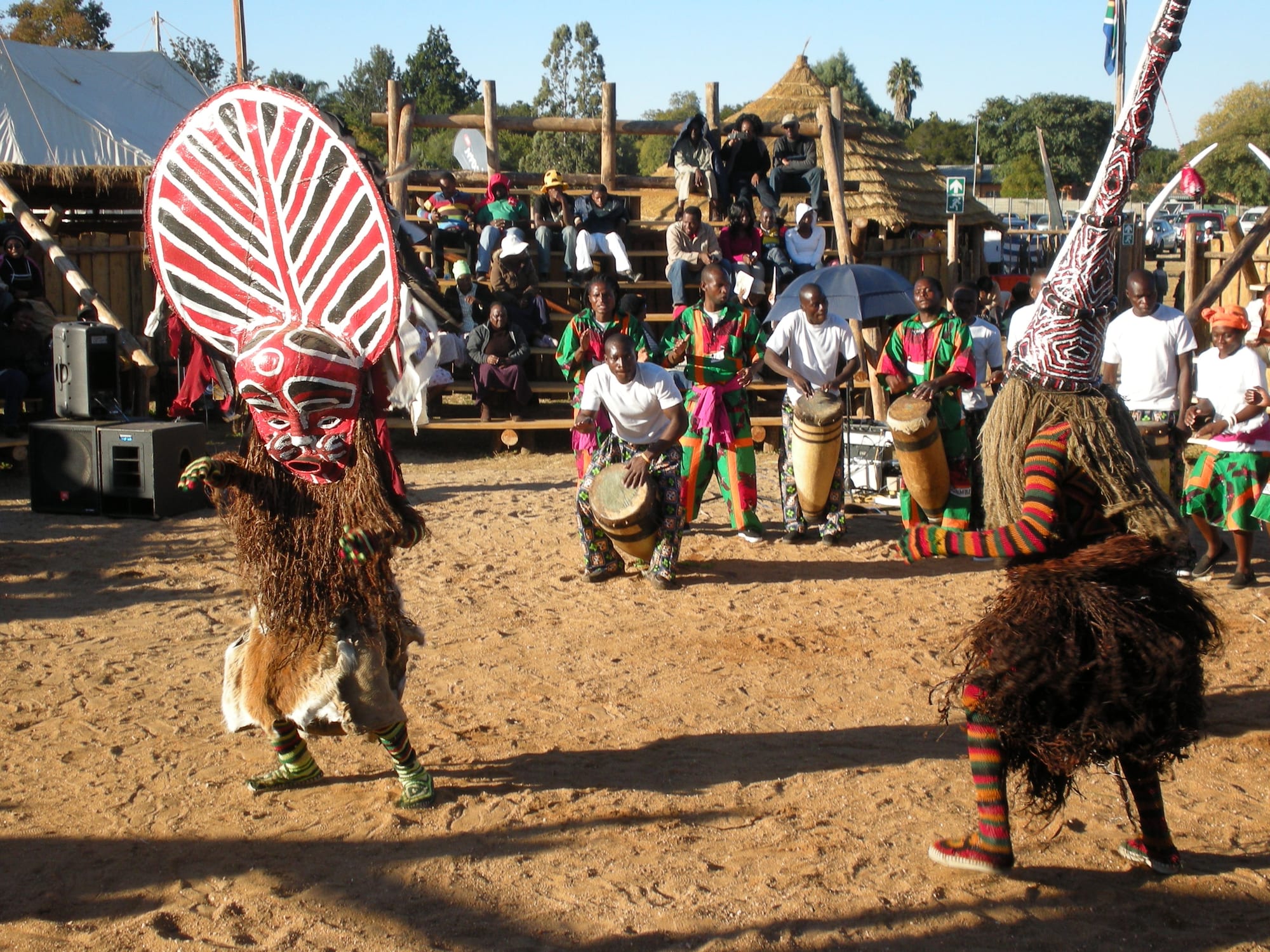 Two masked dancers in fibre costumes and striped leggings perform on sandy ground, one wearing a large red and white striped mask, while drummers play behind them and spectators watch from wooden stands