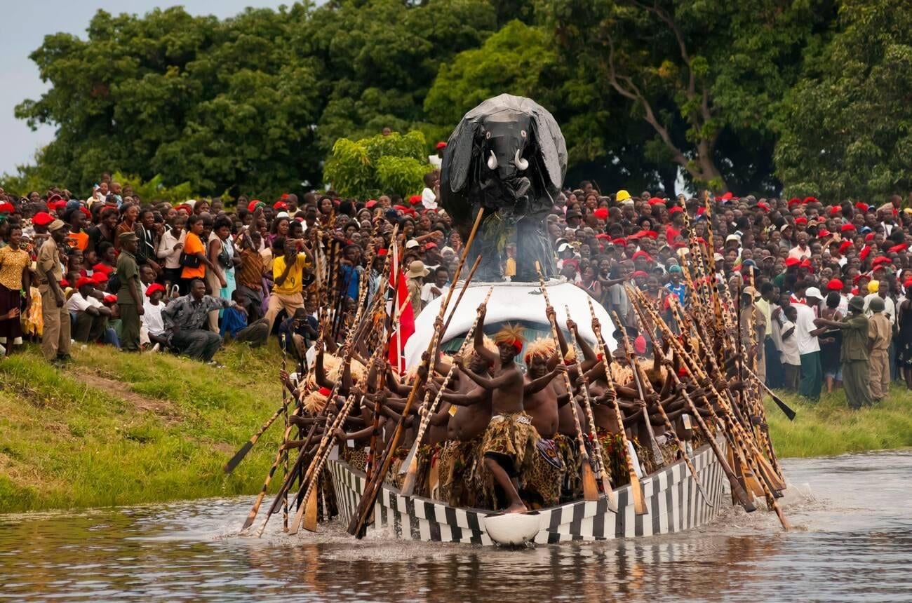 A long black-and-white striped barge topped by a large black elephant figure is driven through the water by rows of paddlers in grass skirts, with a dense crowd in red berets lining the bank behind
