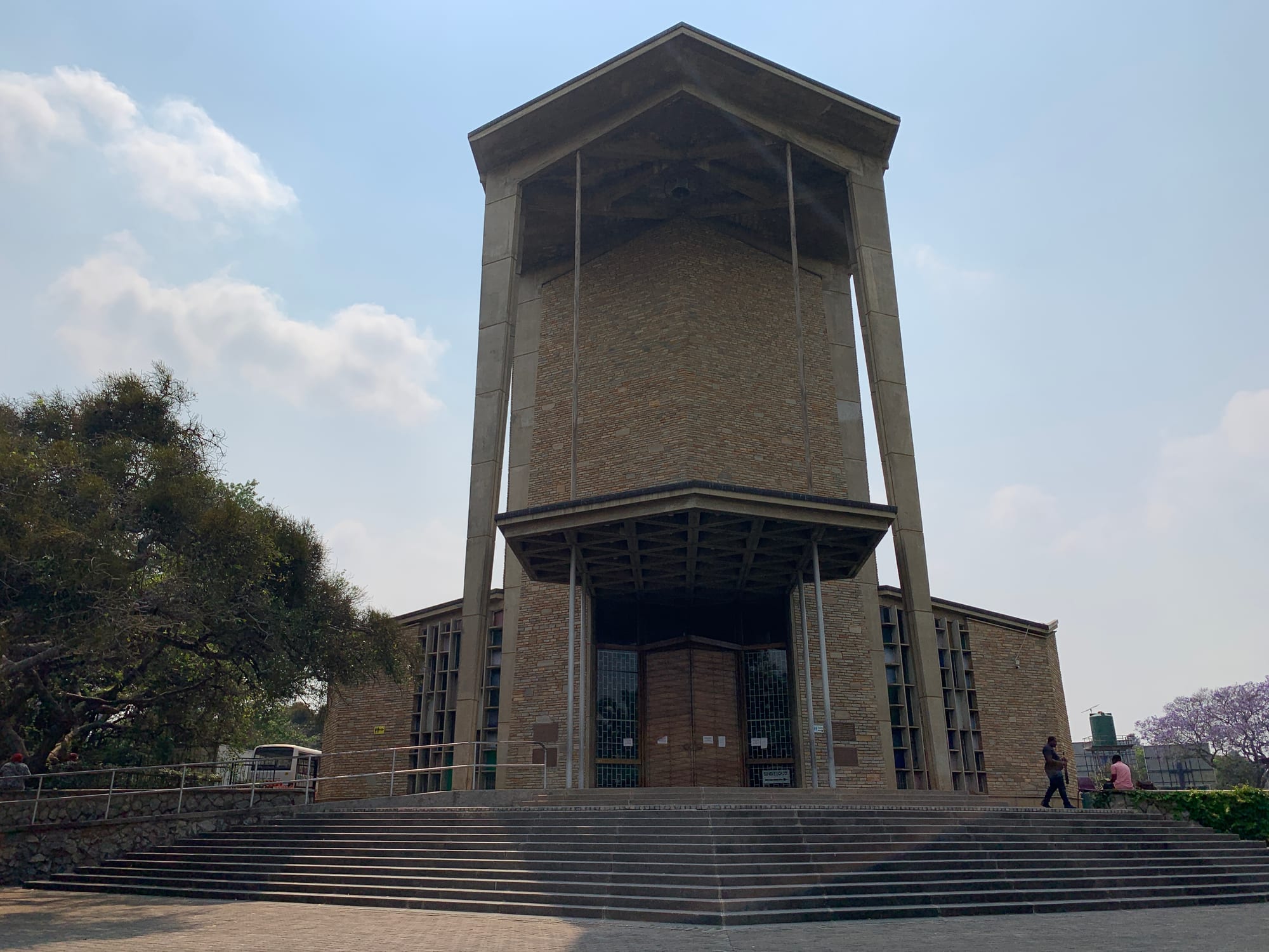 A tall, modernist church of pale stone with a projecting roof and entrance canopy, reached by a wide flight of steps, under a hazy blue sky
