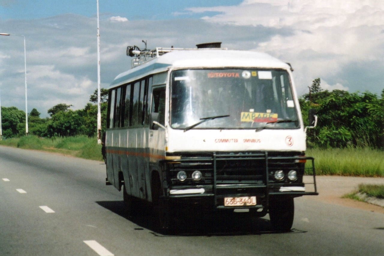 A white and blue commuter minibus marked Commuter Omnibus, driving along a road bordered by grass and trees