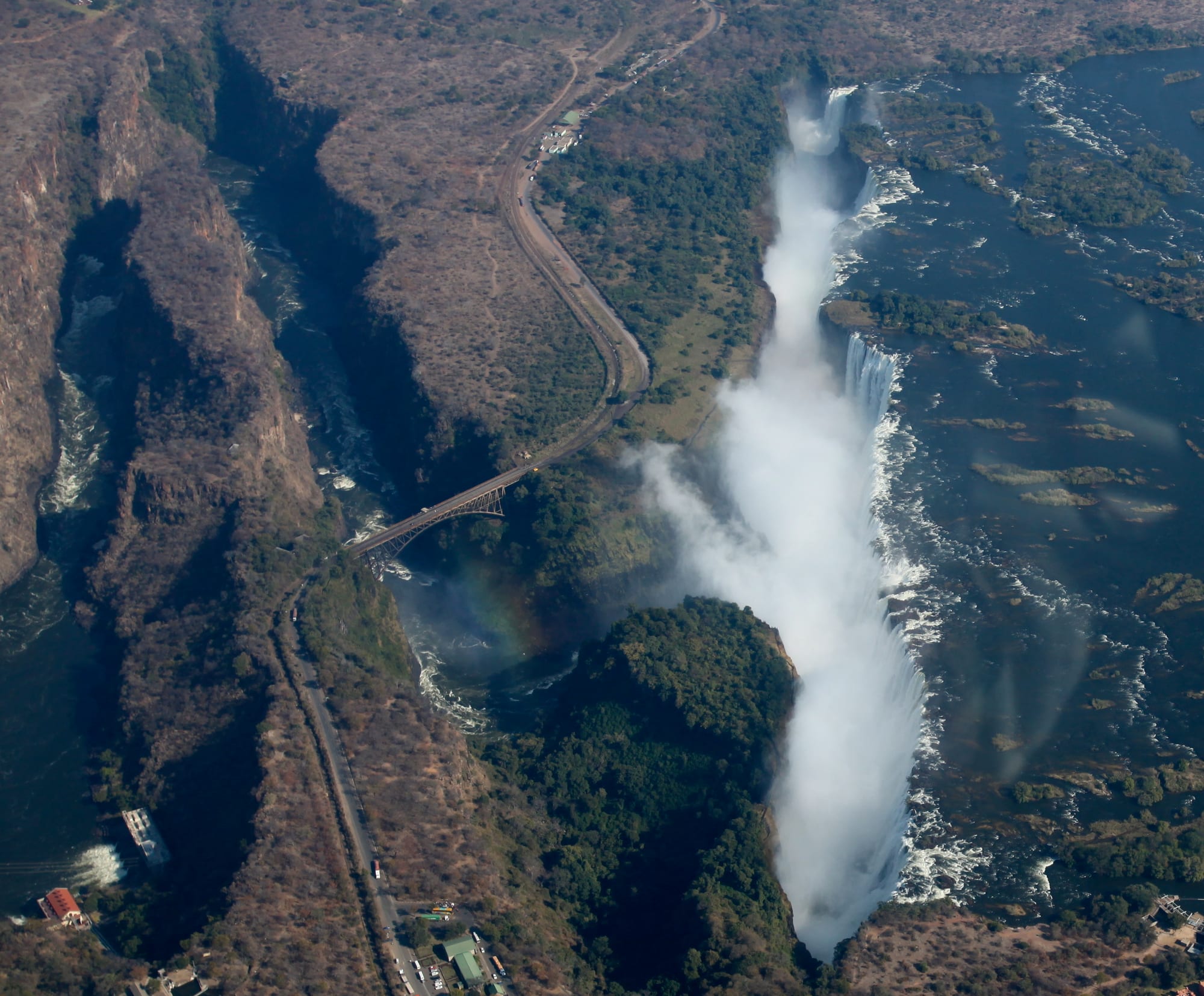 A direct aerial view of a waterfall pouring into a narrow gorge, with a bridge and rainbow visible below