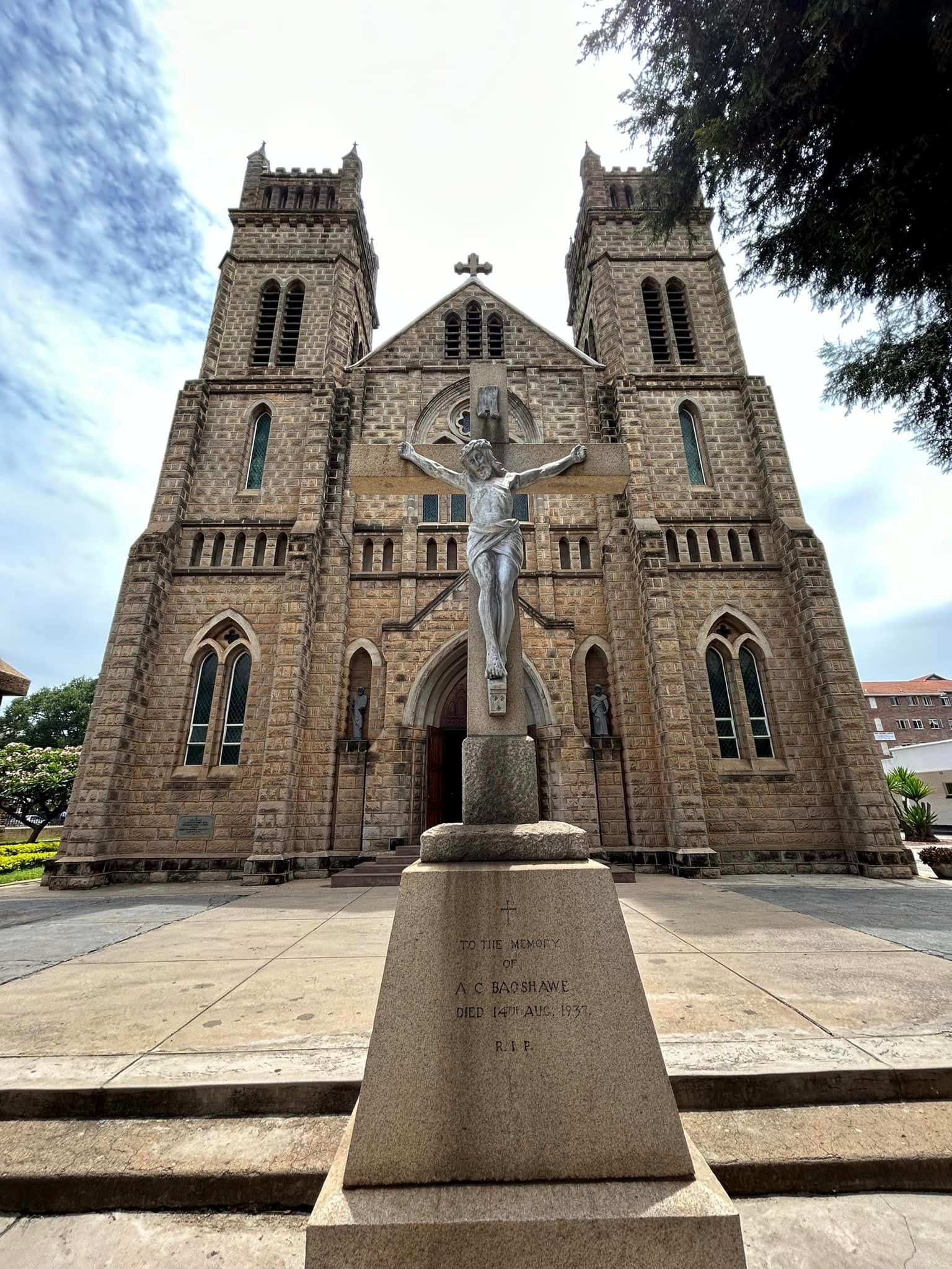 The twin-towered stone facade of a Gothic-style cathedral, with a large crucifix on a stone plinth in front of the entrance