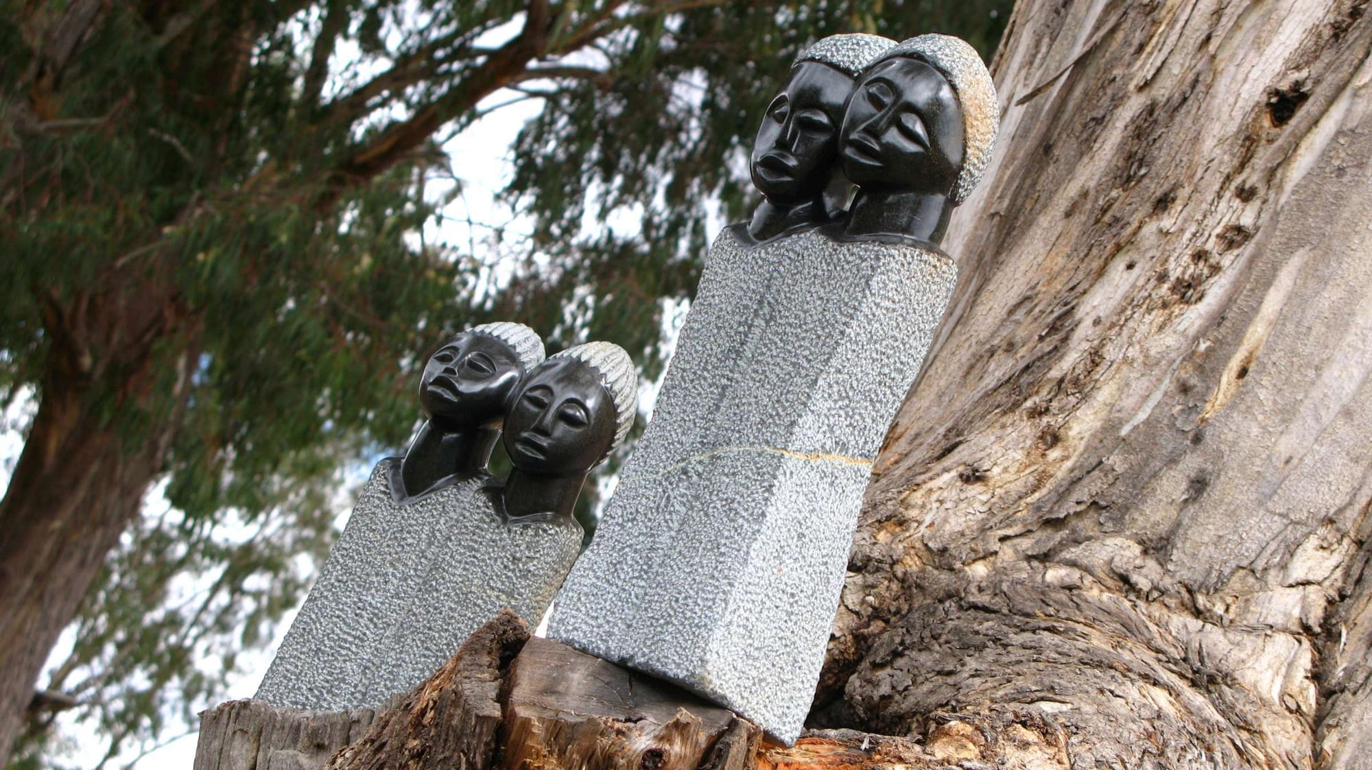 Two stone sculptures, each of two stylised heads with polished dark faces rising from rough-textured grey stone bases, set on a tree stump