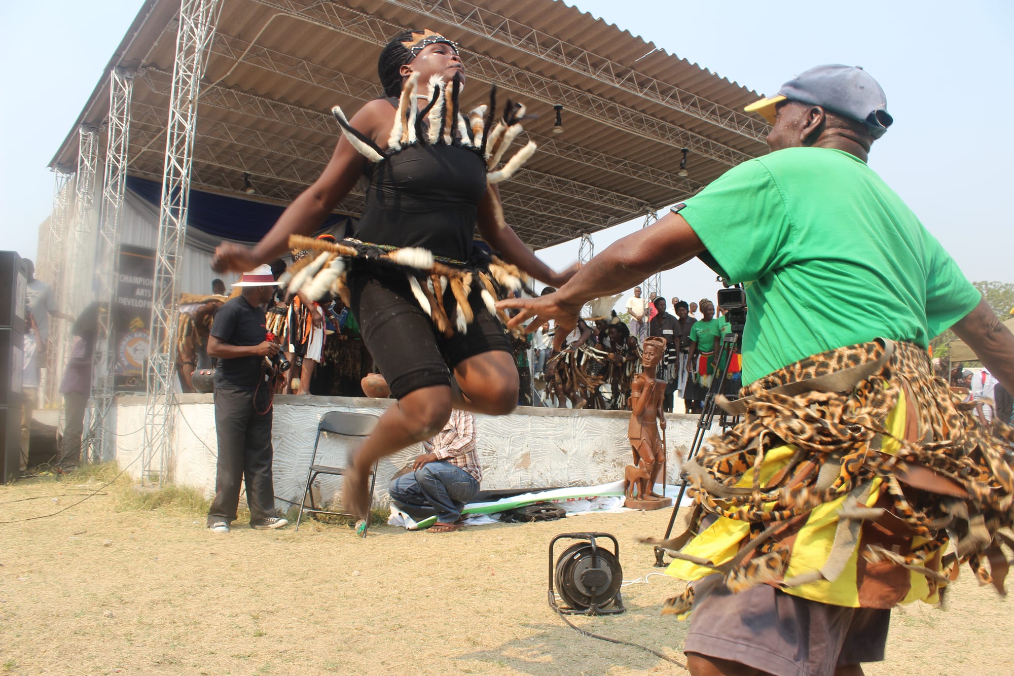 A woman in a black costume with white fringed decorations leaps high off the ground as an older man in a green shirt and leopard-print wrap dances beside her, with a covered stage and onlookers behind