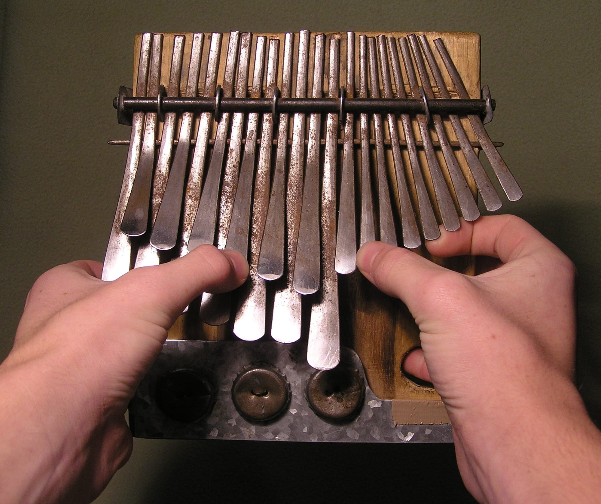 Two hands holding a wooden mbira with two rows of flattened metal keys, the thumbs resting on the keys and bottle-cap rattles fixed to a metal plate at the base