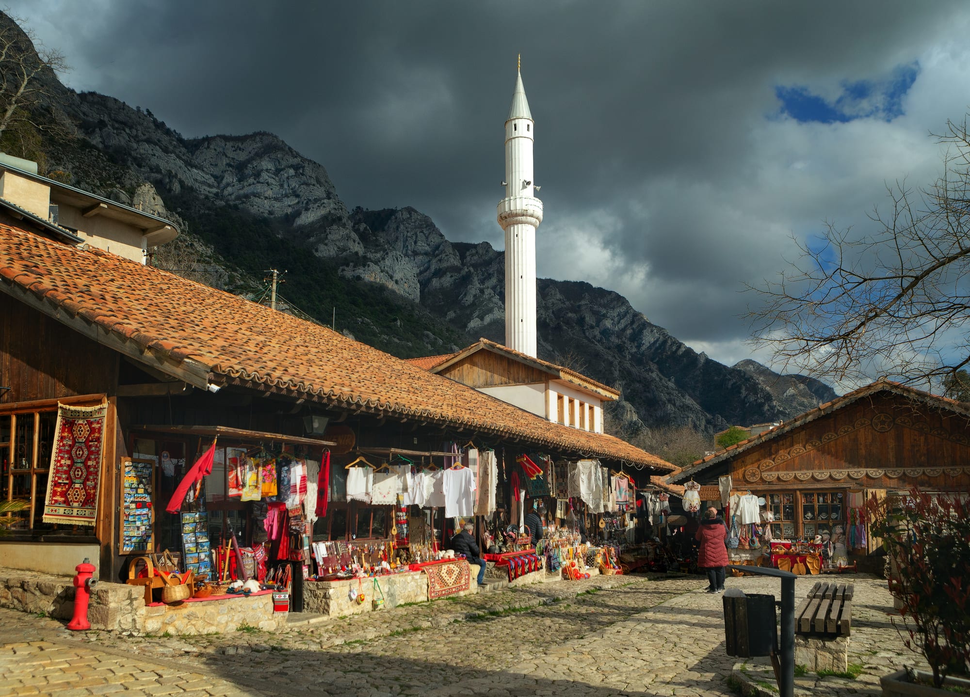 Wooden bazaar shops hung with rugs and textiles on a cobbled street, a white minaret and grey mountains behind