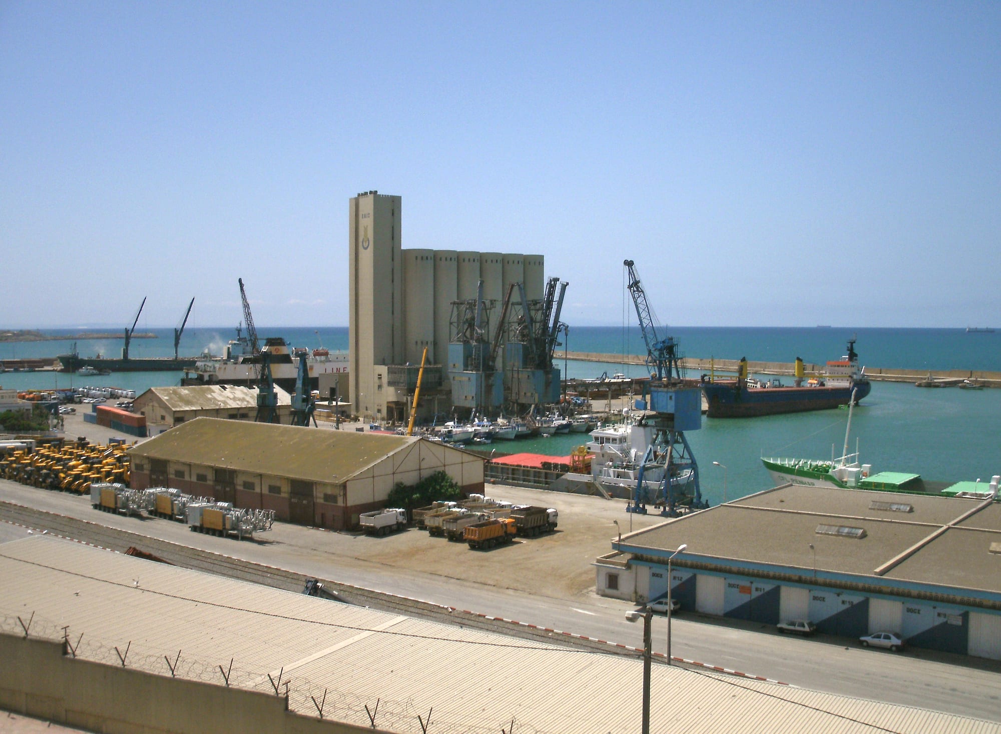 Cranes, grain silos and cargo ships in a harbour beside turquoise sea, with warehouses and lorries on the quays below