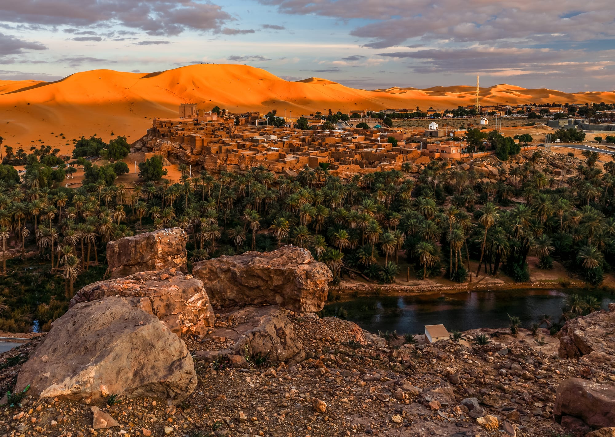An earthen village on a rocky rise above a dense palm grove and a narrow watercourse, with a long orange dune ridge rising behind it
