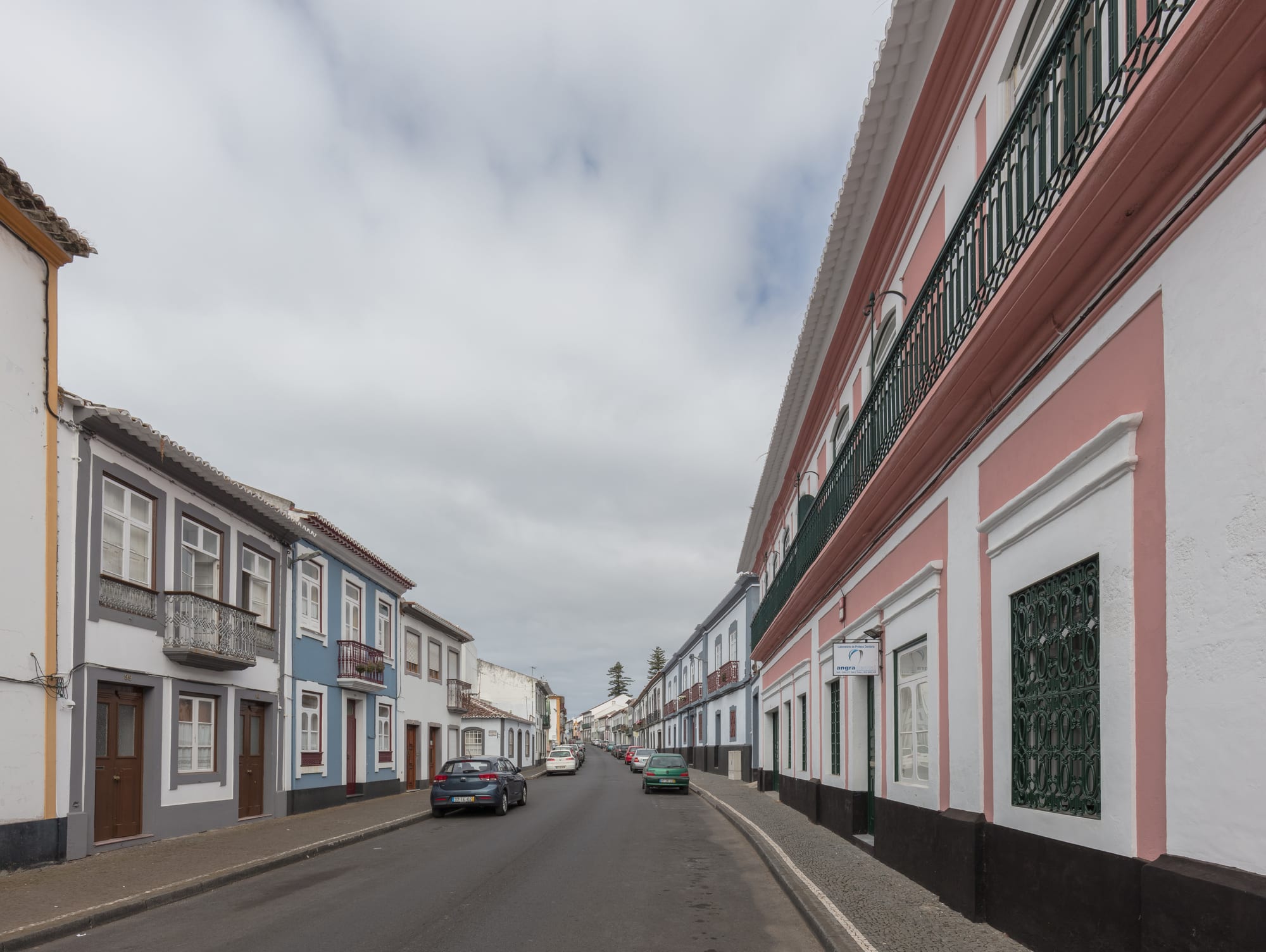 A straight street of two-storey houses painted white, pink, blue and grey, with iron balconies and dark stone bases