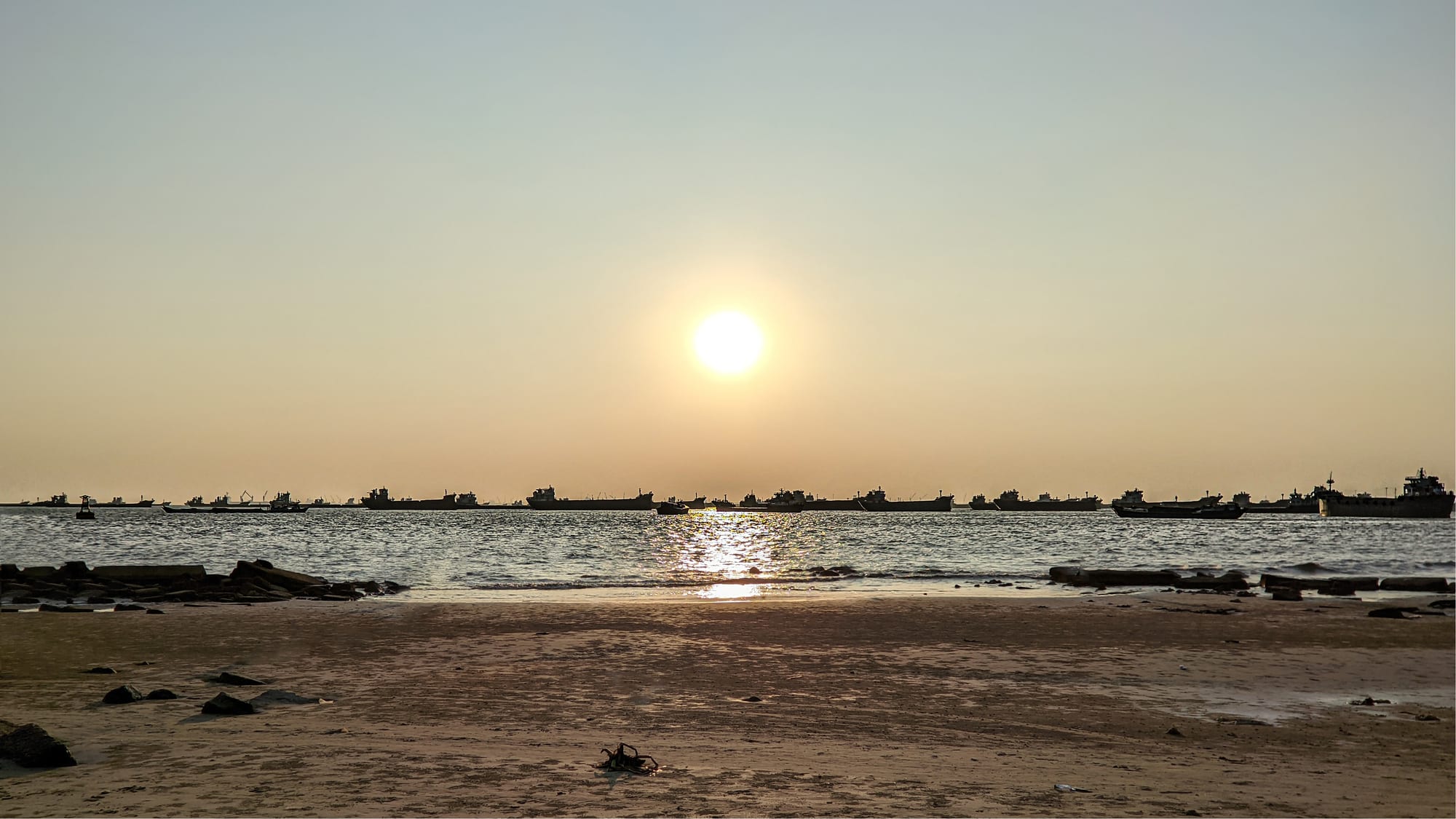 The sun low over the Bay of Bengal at Patenga, with a line of anchored cargo ships on the horizon and wet sand and stones in the foreground