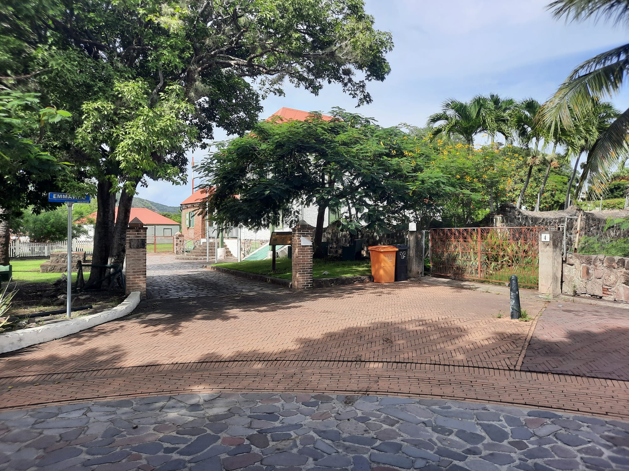 A brick-paved street in Oranjestad's Upper Town with shade trees, stone walls and a red-roofed house behind a gate