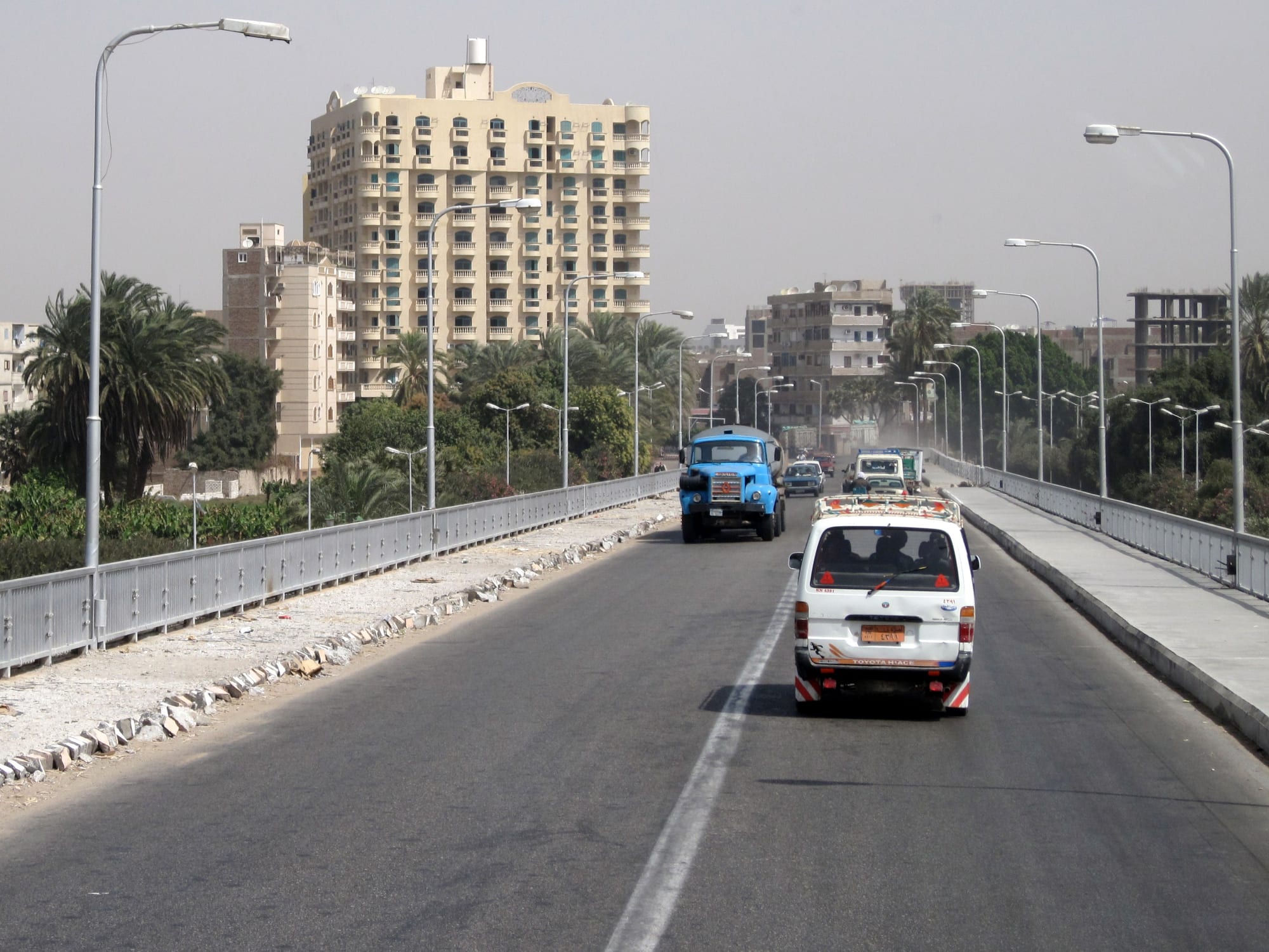 A road bridge over the Nile at Qena with a minibus and lorry crossing, palm trees and pale apartment towers of the city beyond