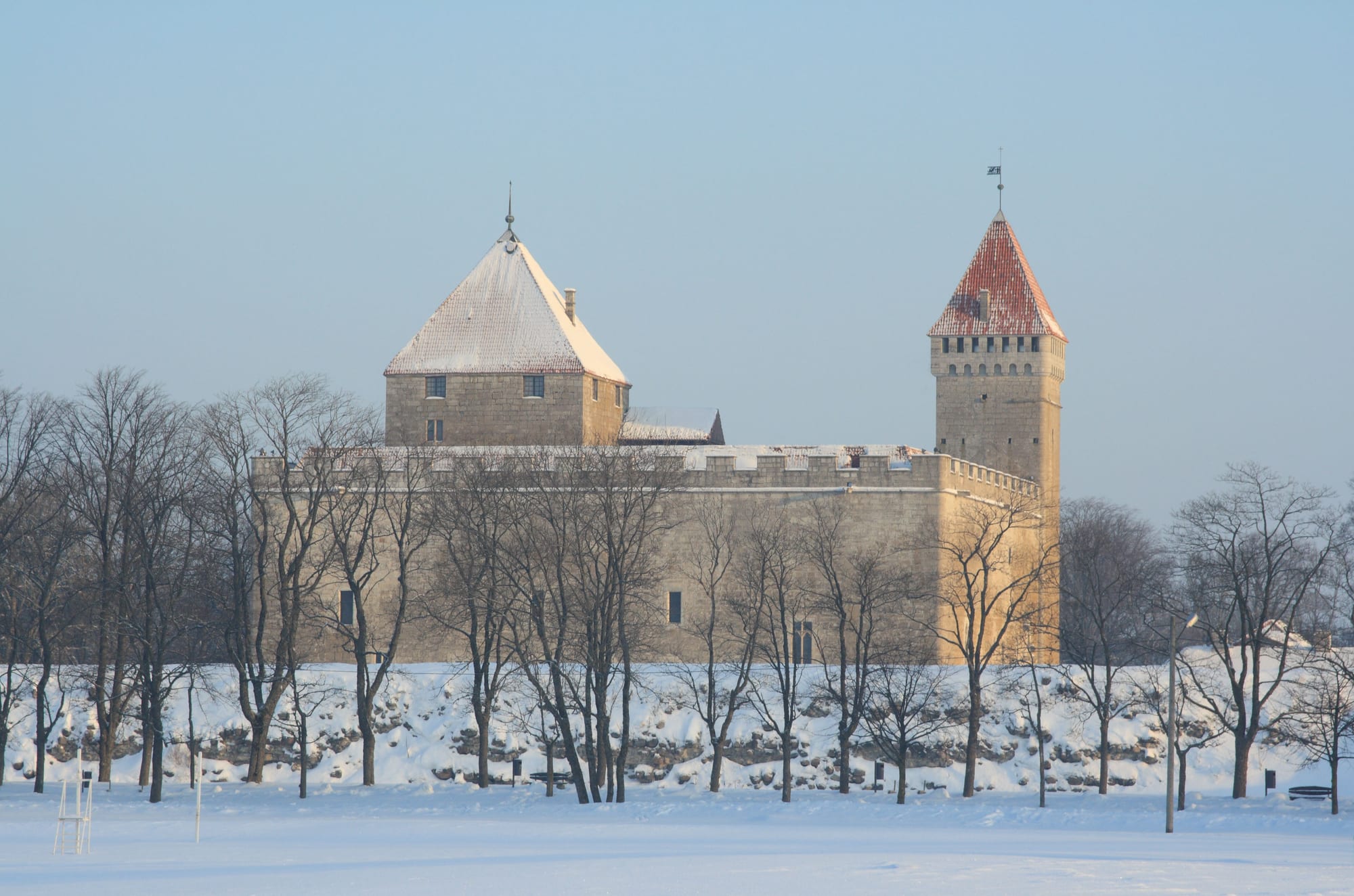 Pale stone walls and a tall square tower with a red pointed roof behind bare trees and a snow-covered moat bank