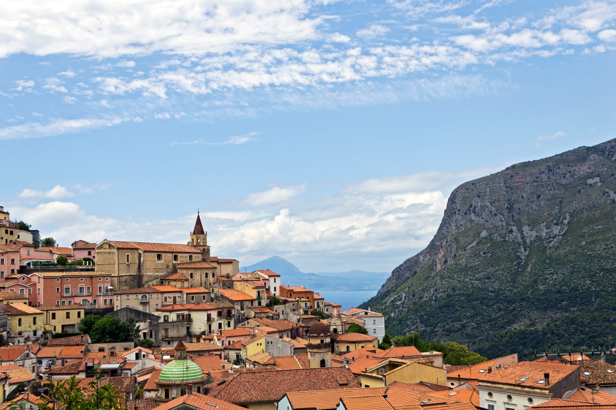 Pastel houses and tiled roofs of a hill town with a pointed bell tower and a green-tiled dome, beside a steep wooded mountain