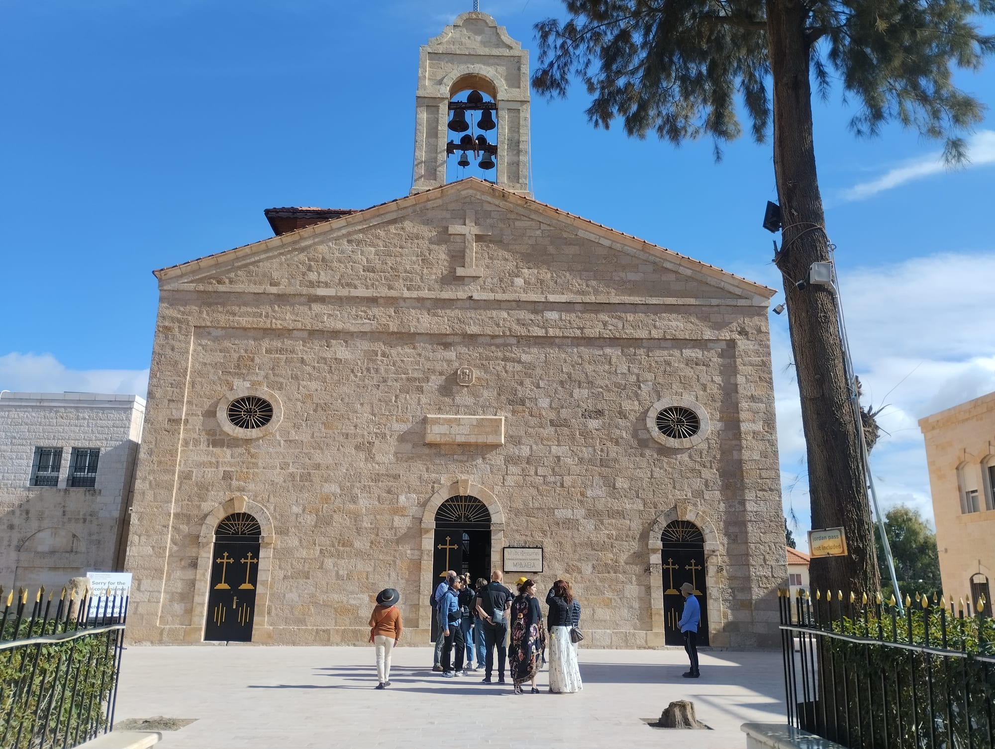 The pale stone west front of St George's Church in Madaba, with an open bell arch above the gable and a pine tree to one side