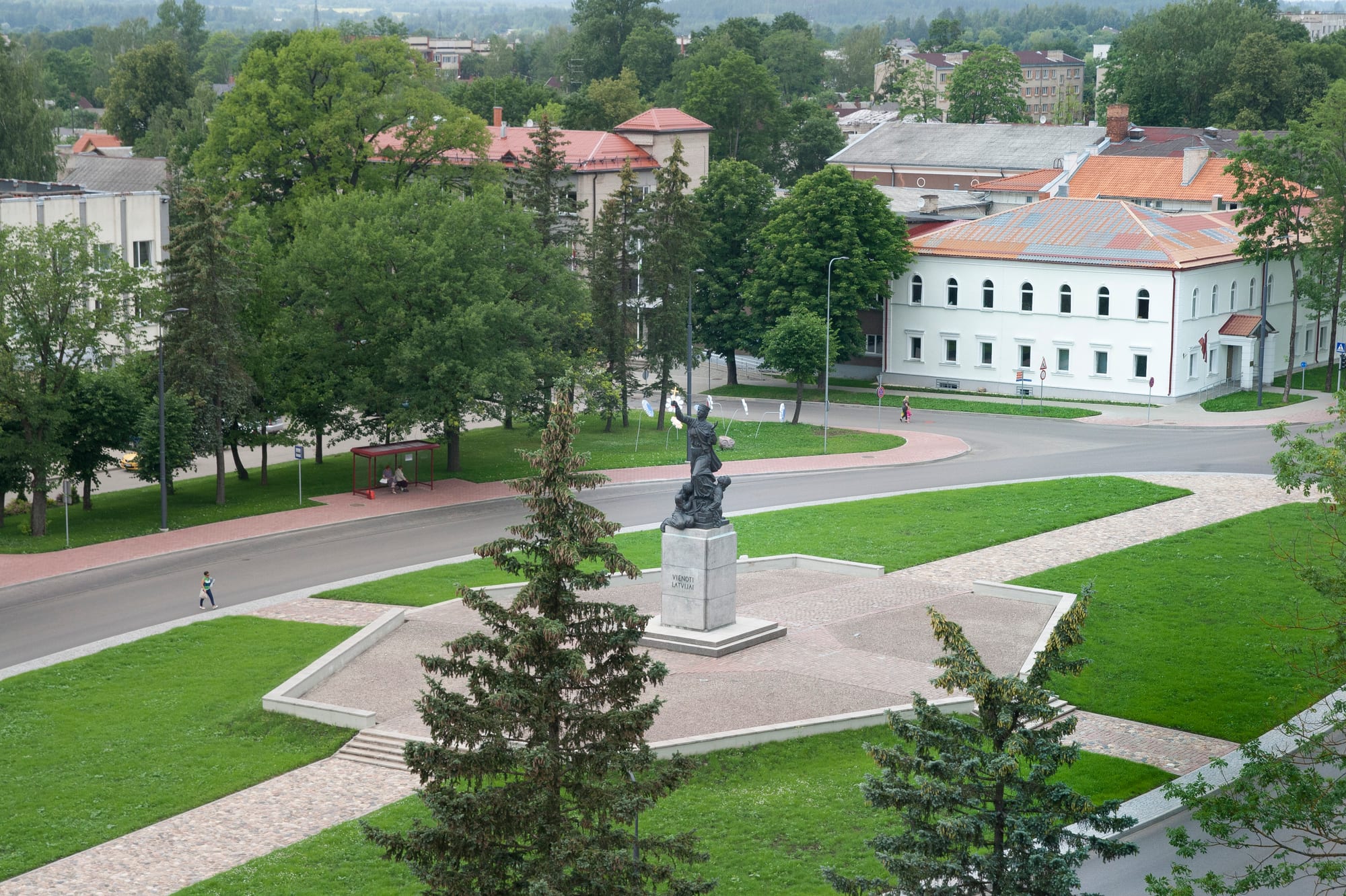 A bronze statue on a stone plinth in a green square beside a road, with trees and low red-roofed buildings behind
