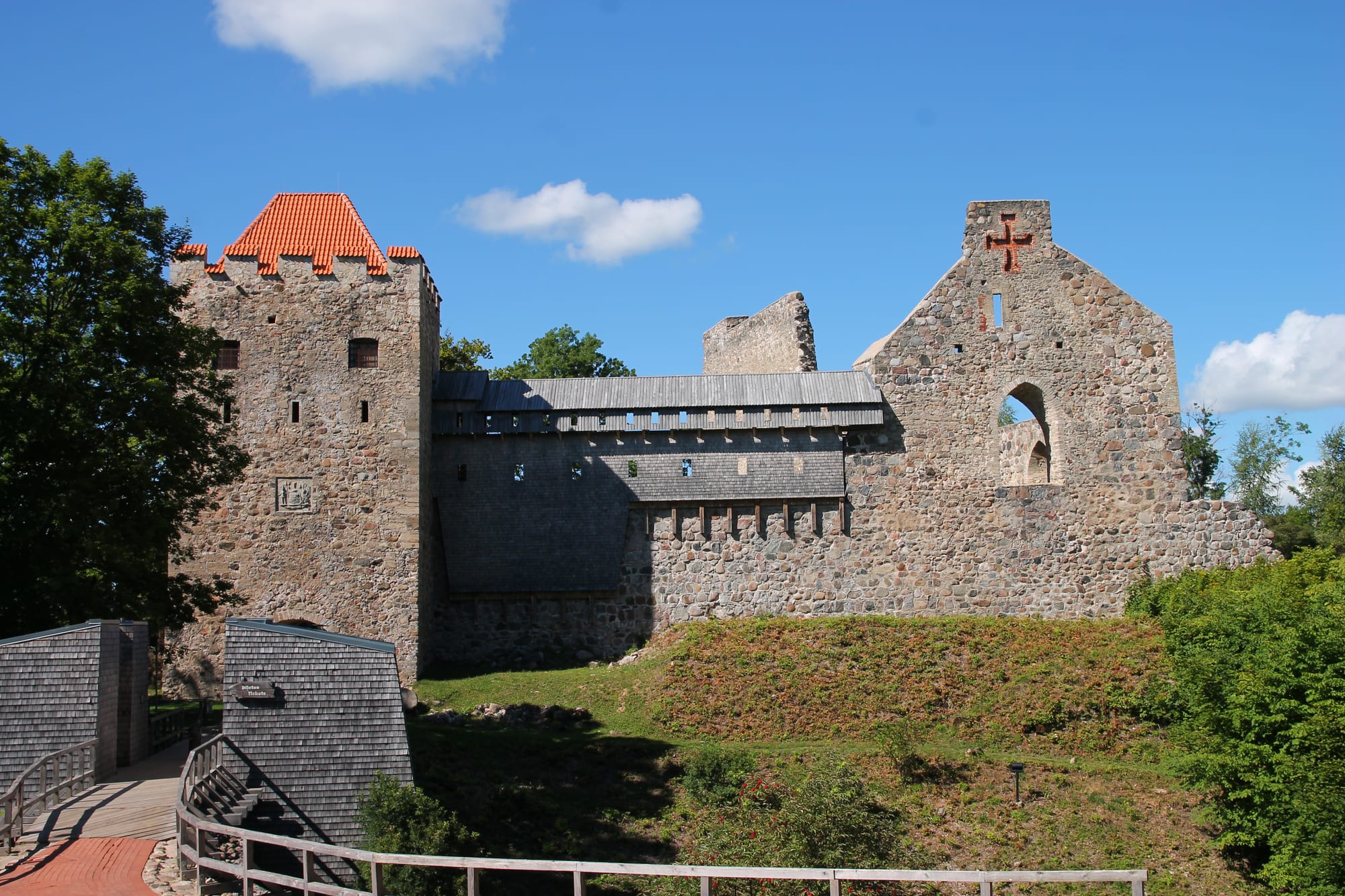 Stone walls and a red-roofed gate tower of Sigulda's medieval castle ruins under a blue sky, with a wooden walkway in front