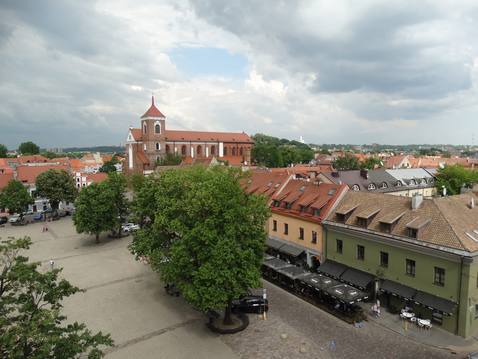A cobbled old-town square with linden trees and café awnings, red-tiled roofs and a brick cathedral beyond