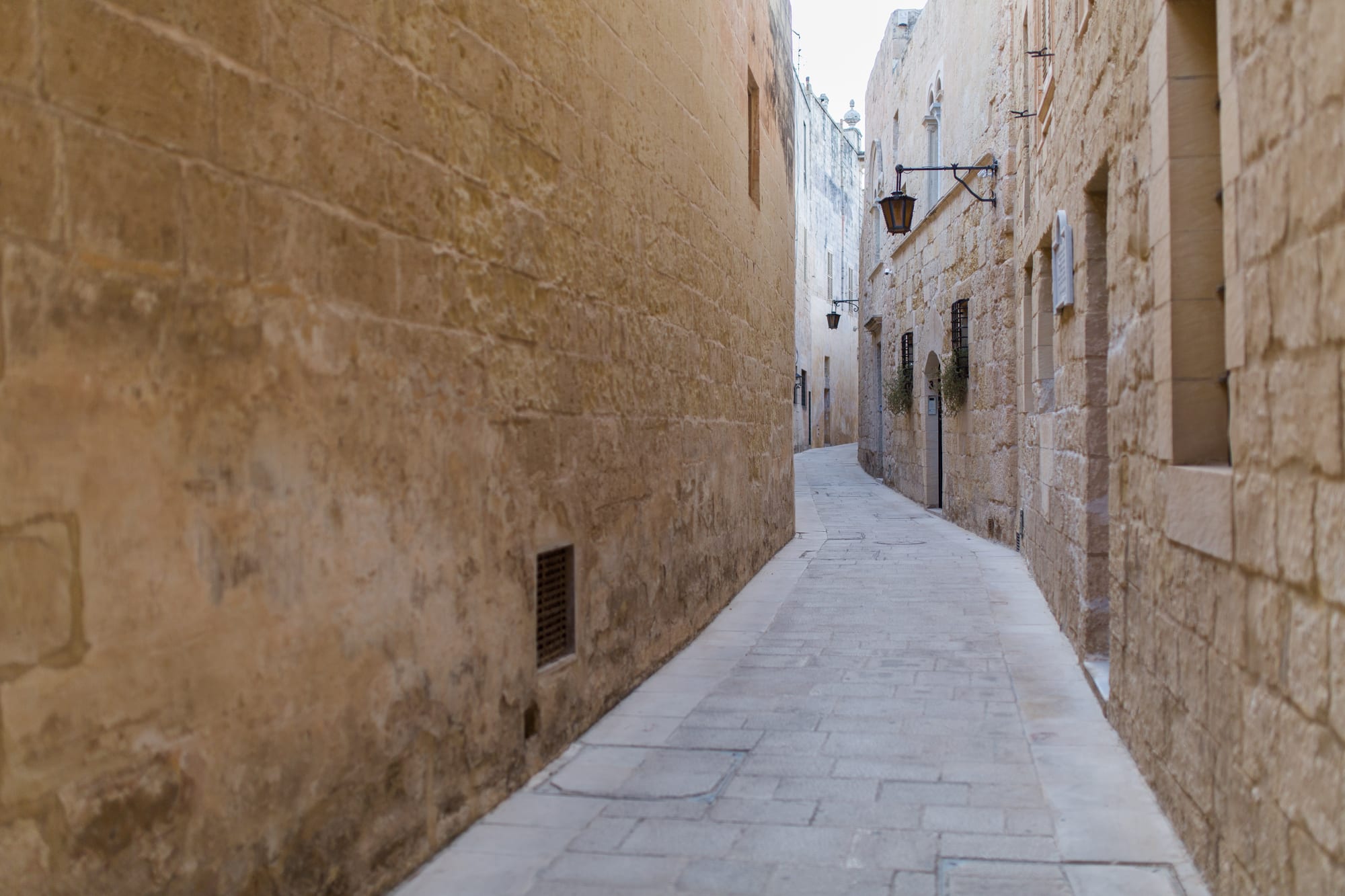 A narrow, empty lane in Mdina between tall honey-coloured limestone walls, with iron lanterns on the buildings