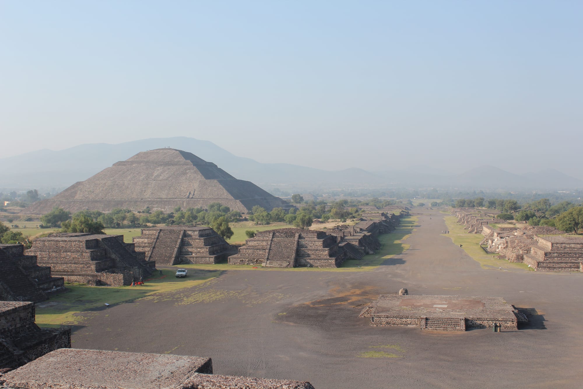 The broad Avenue of the Dead stretching south past stepped platforms, with the Pyramid of the Sun on the left and hazy hills beyond