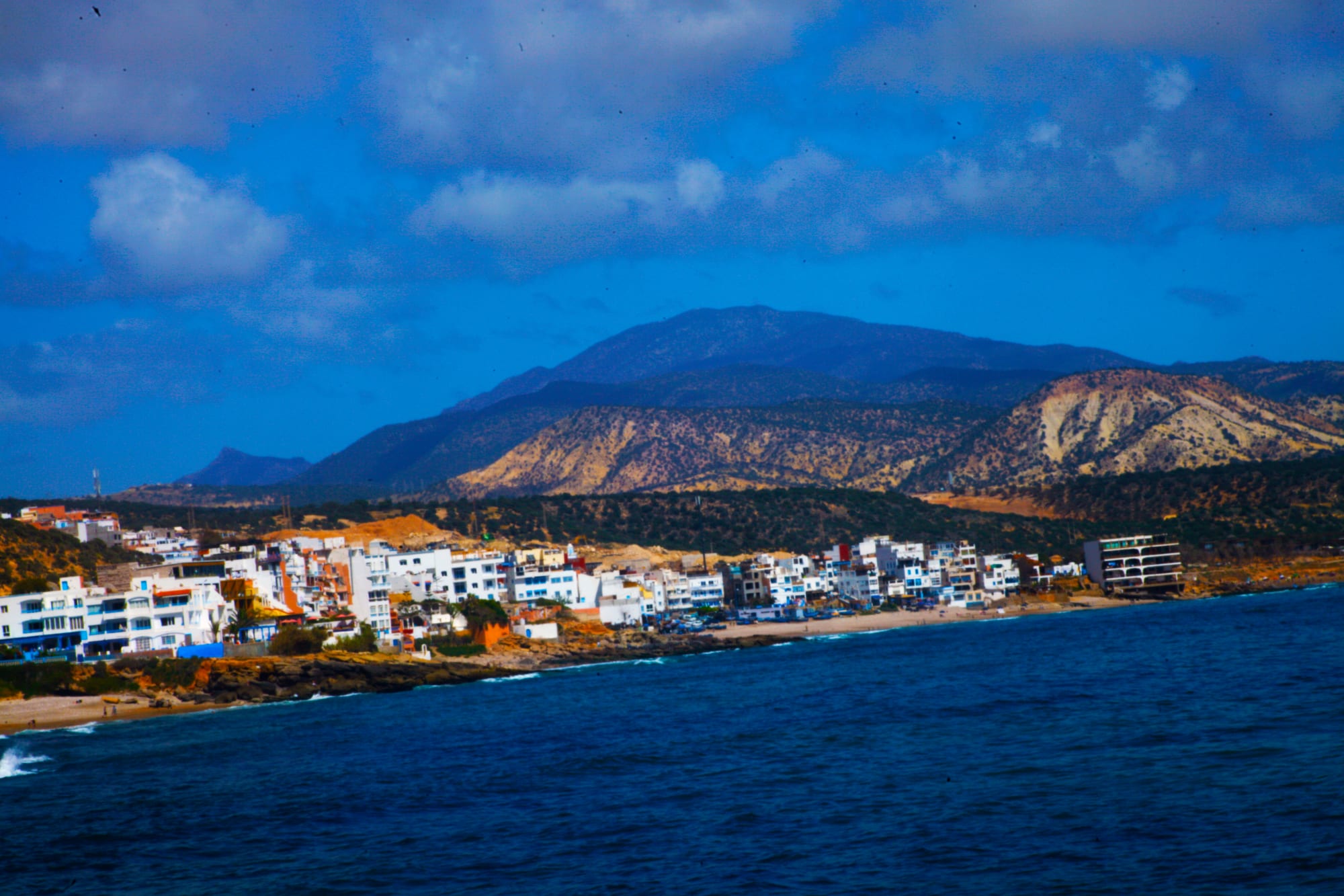 A cluster of white and pale buildings on a rocky Atlantic shore, with dark hills rising behind and surf breaking along the coast