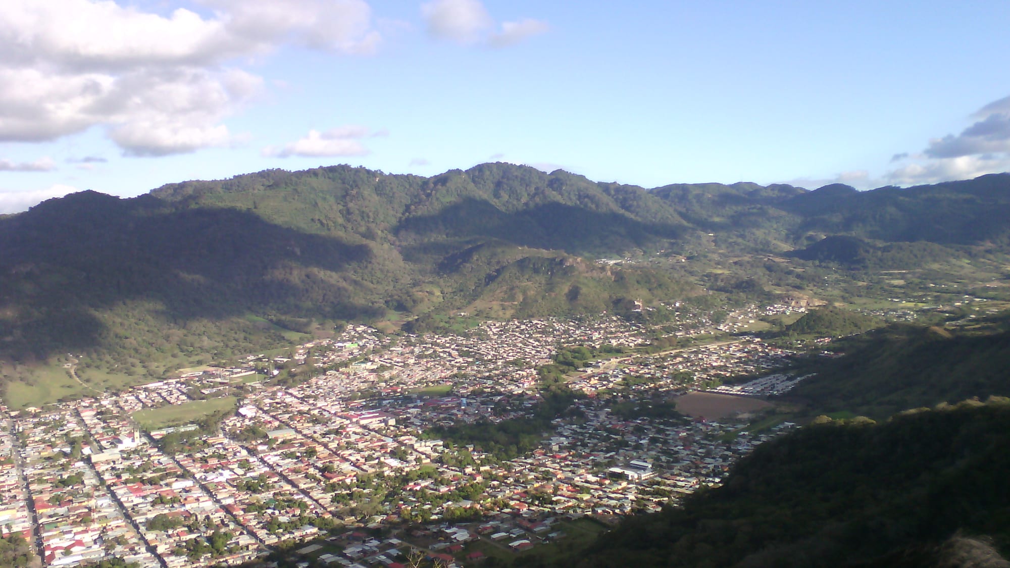 A town of red and grey roofs spread across a green valley floor, ringed by forested mountains under a blue sky