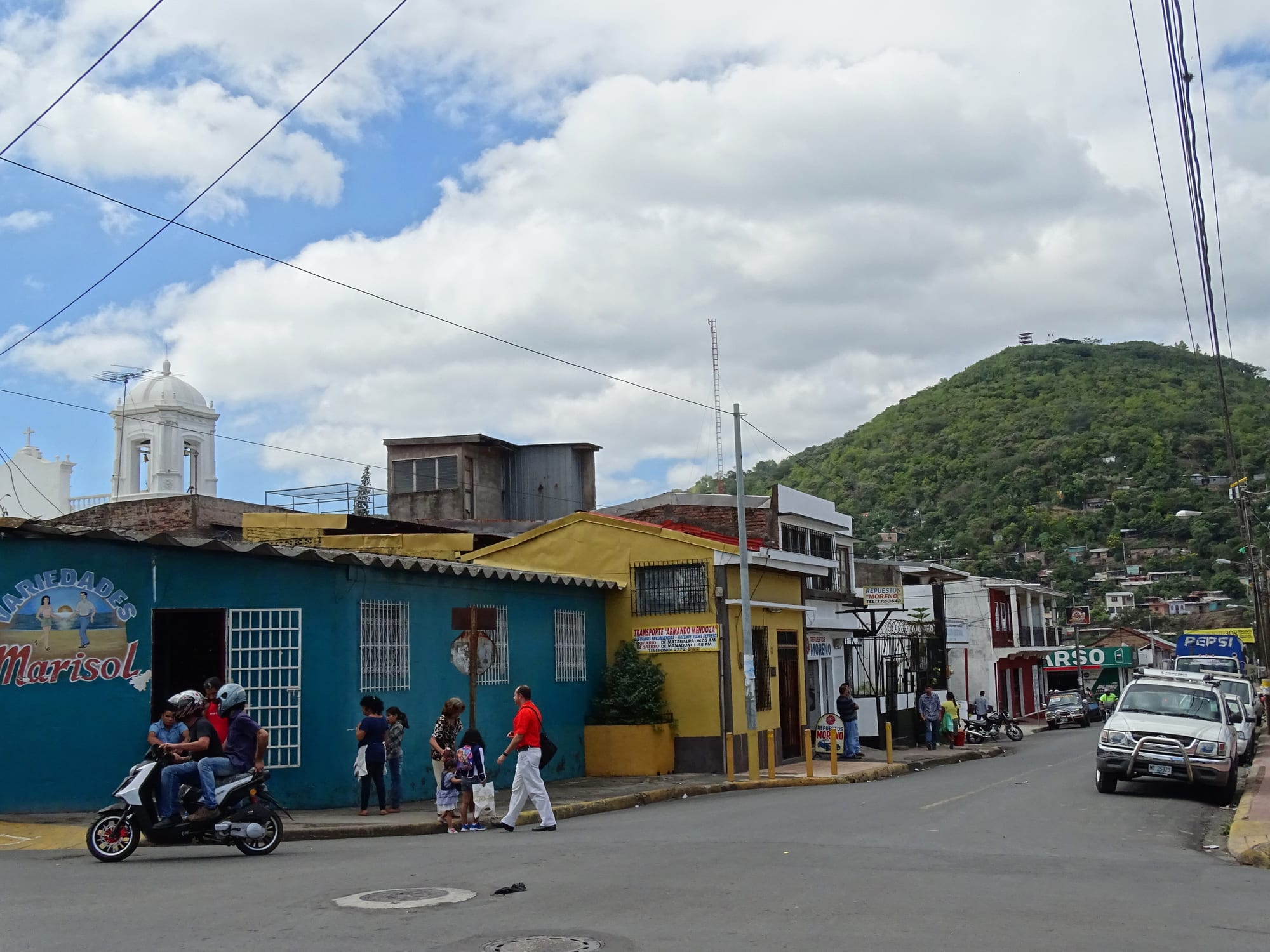 A street corner of low painted shops with a white church bell tower on the left and a green wooded hill rising behind