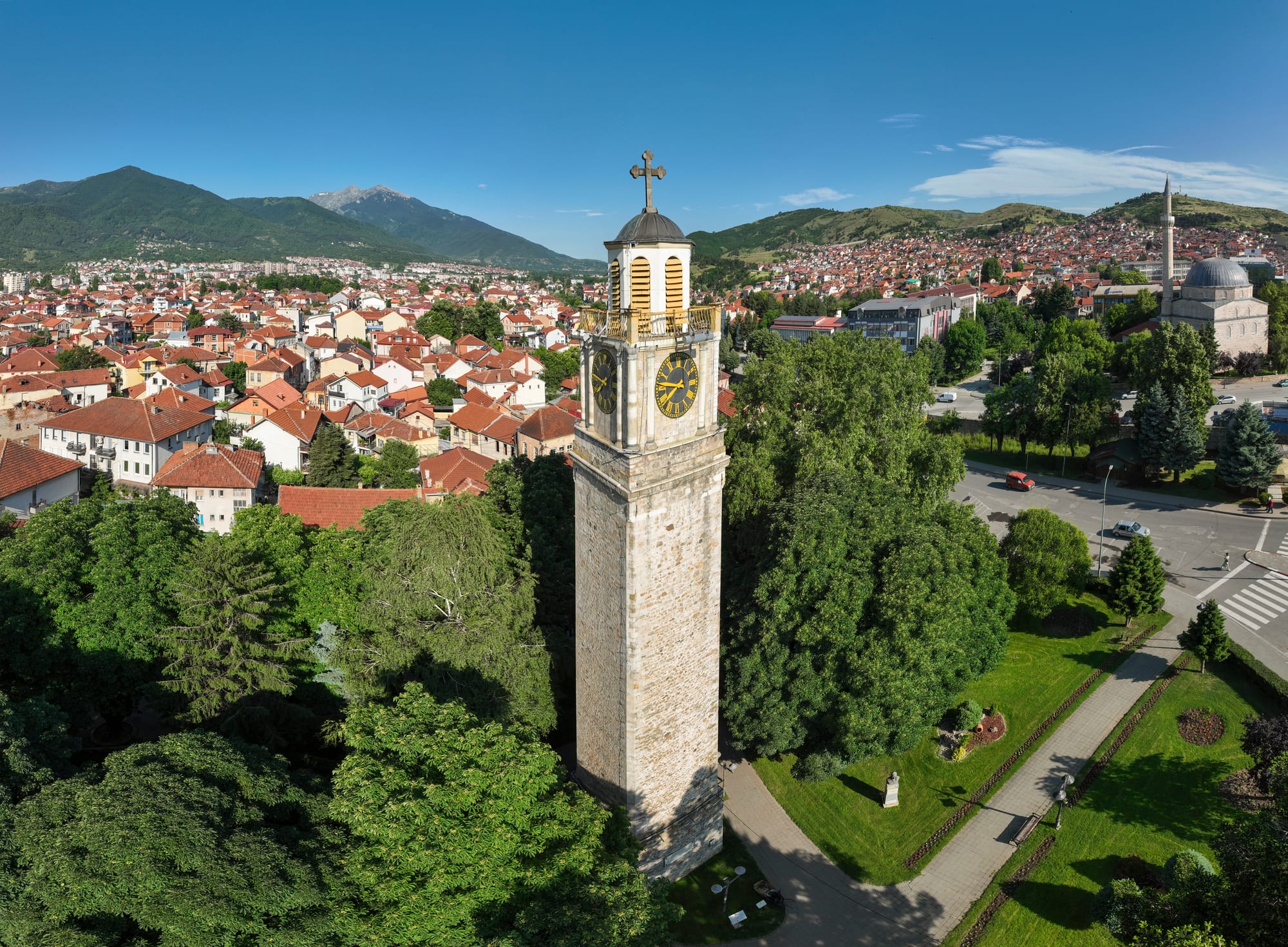 Bitola's stone clock tower rising above trees, with red-roofed houses, a domed mosque and green mountains behind