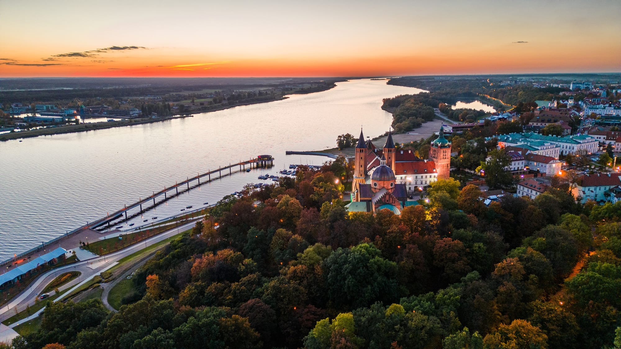 Aerial view at sunset of Płock's brick cathedral and castle among autumn trees above the wide Vistula, with the town to the right