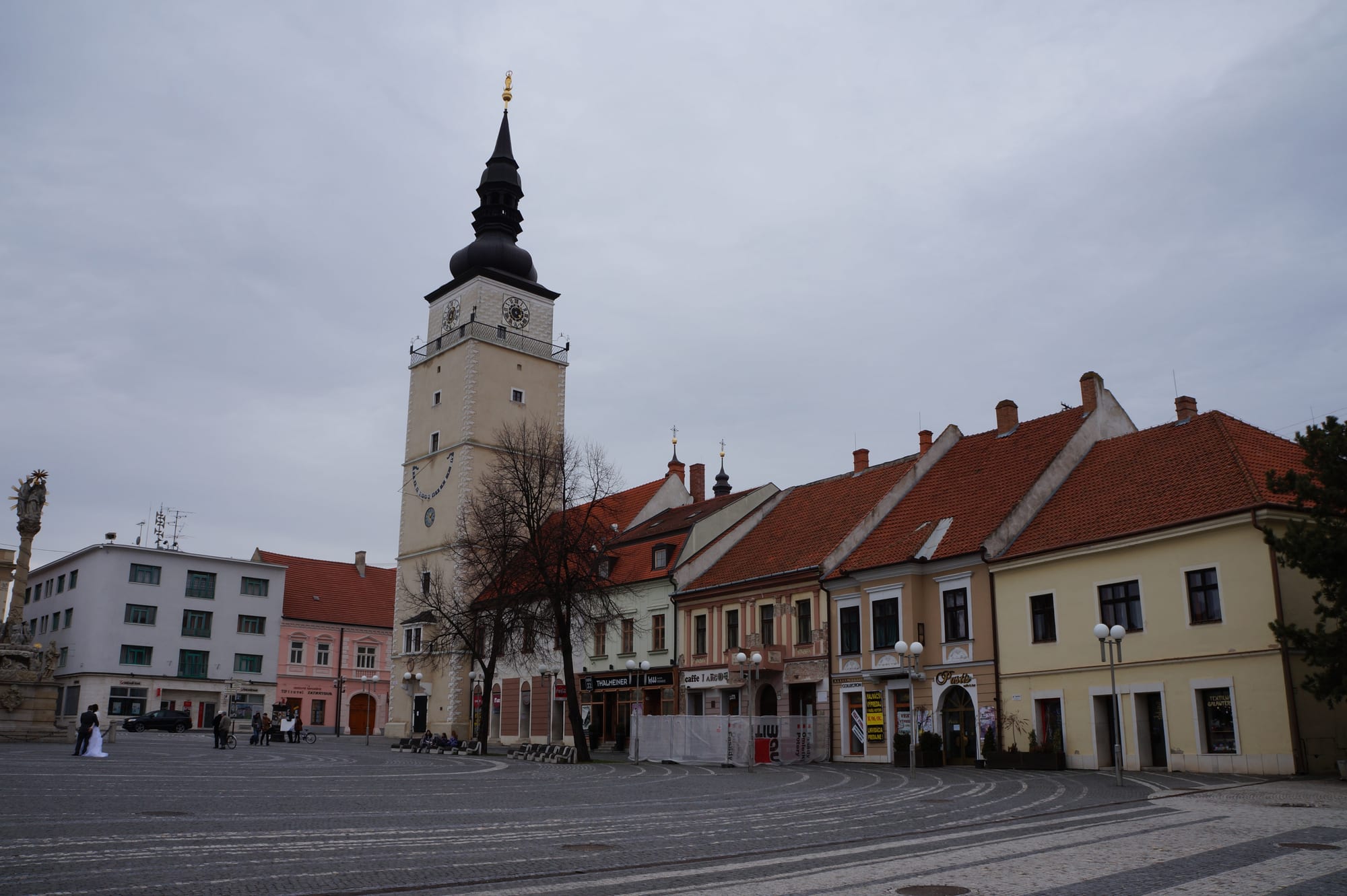 A wide cobbled square with a tall cream clock tower topped by a dark onion spire beside a row of red-roofed pastel houses