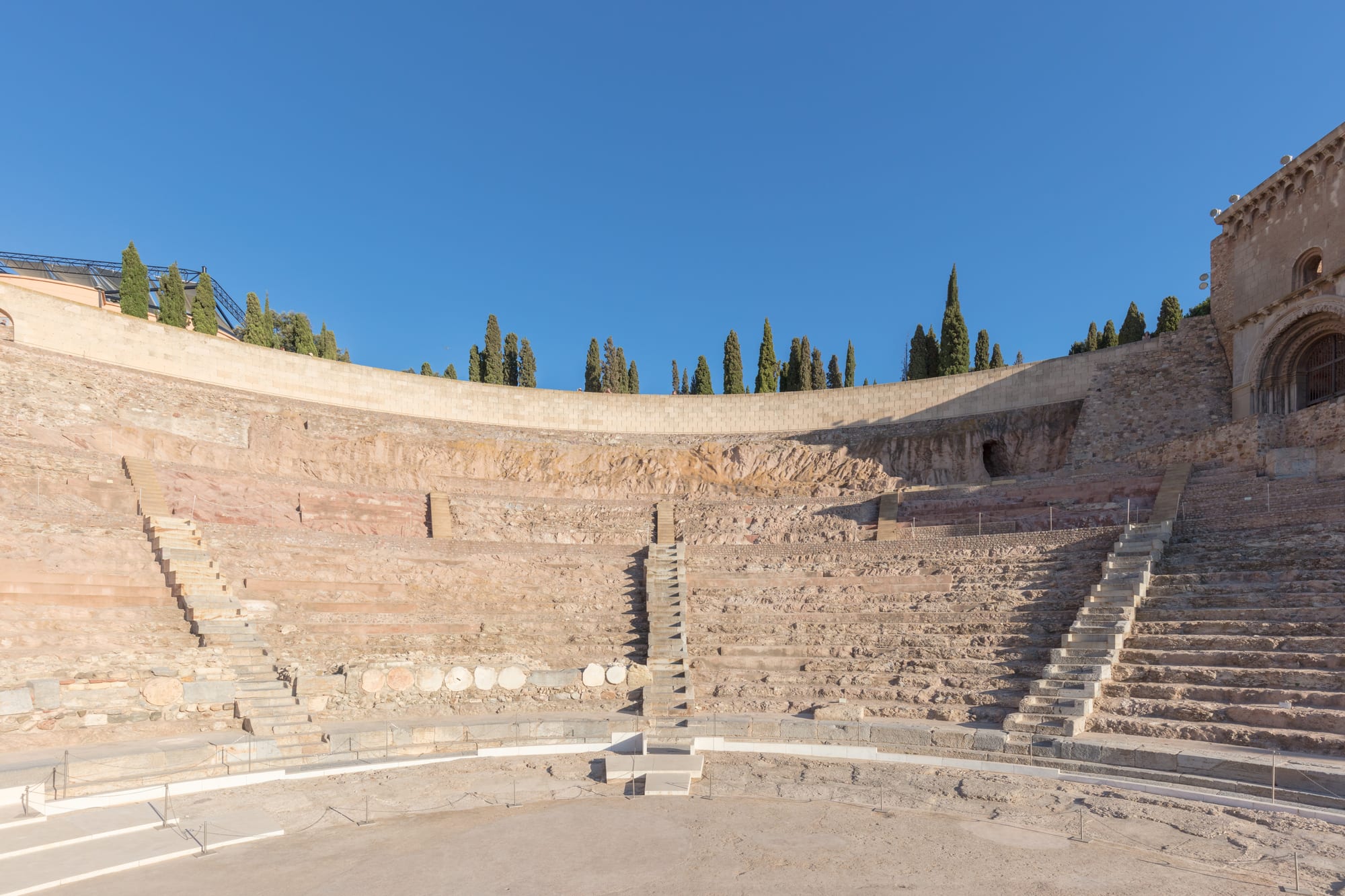 The curved stone tiers and stairways of a restored Roman theatre, with cypress trees and a ruined church wall above under a clear blue sky